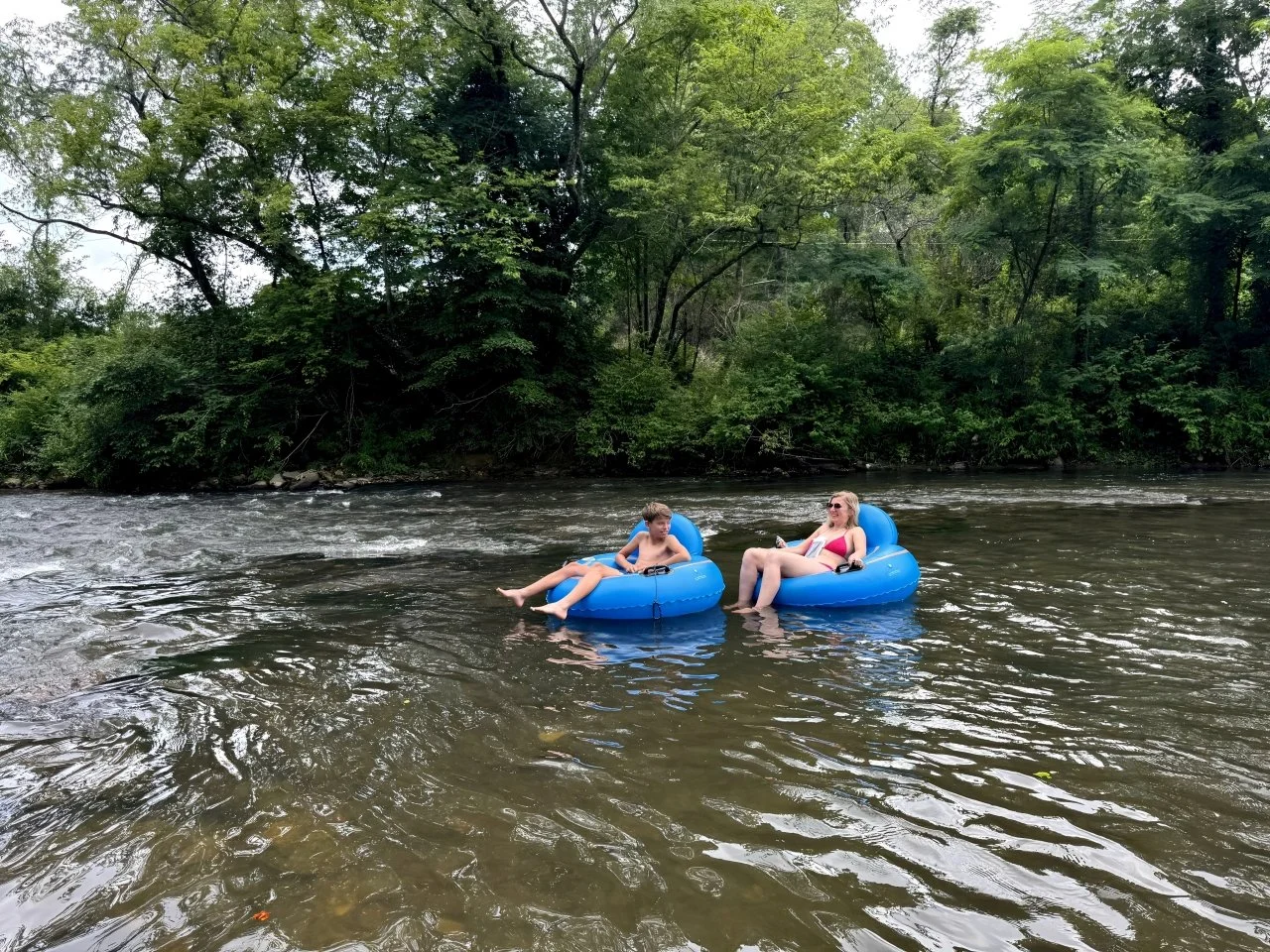 Two people, a man and a woman, relaxing on blue float tubes in a river surrounded by dense green trees.