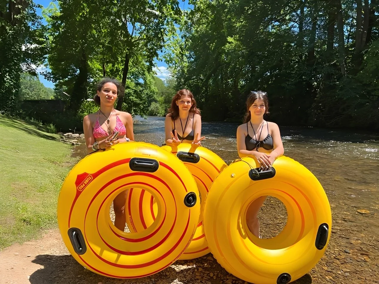 Three young women in swimsuits standing on a riverbank, holding yellow inflatable rings, with lush green trees and river in the background, enjoying a sunny day.