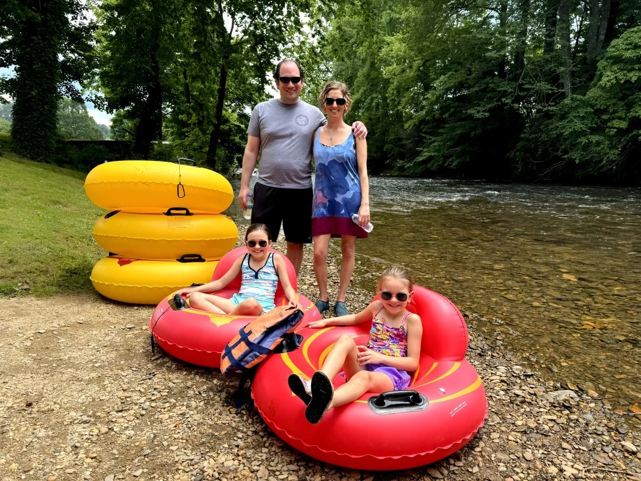 A family of four at the riverbank with yellow and red inflatable tubes, smiling and wearing sunglasses, surrounded by green trees.