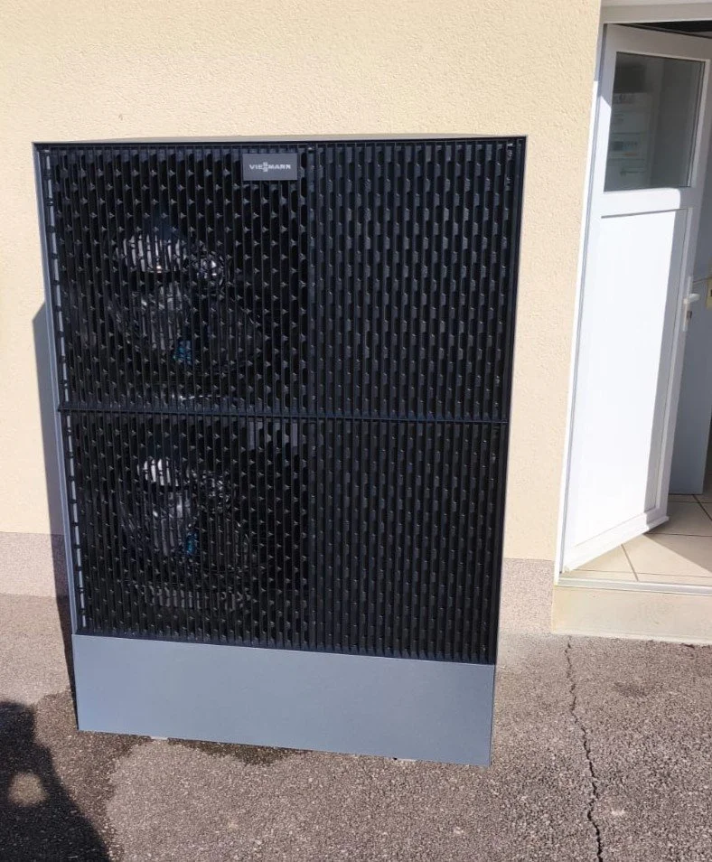 Un climatiseur ou un système de ventilation avec deux ventilateurs derrière une grille noire, placé à l'extérieur d'un bâtiment.