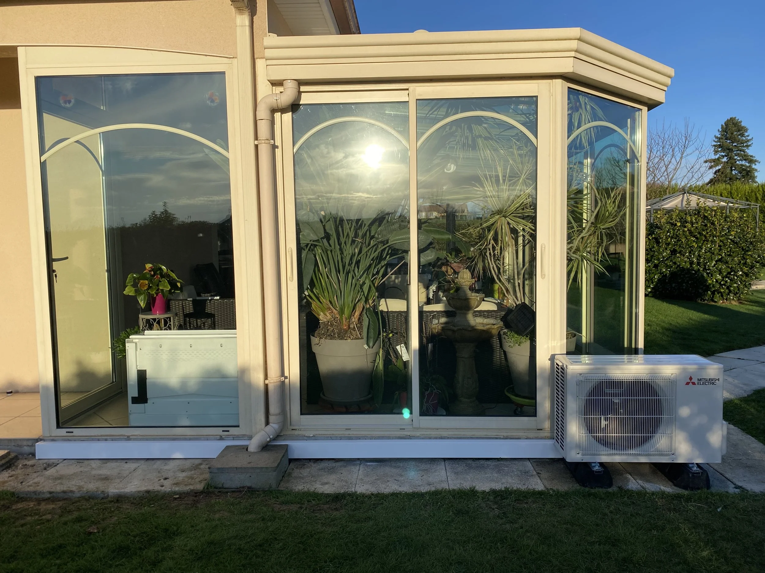 Serre en verre avec plantes d'intérieur, installation de climatisation Mitsubishi Electric, dans un jardin, ensoleillée, avec ciel bleu.