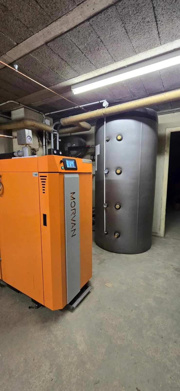 Une installation de chaufferie avec une chaudière orange et un ballon d'eau chaude en acier inoxydable dans une pièce au plafond en béton.