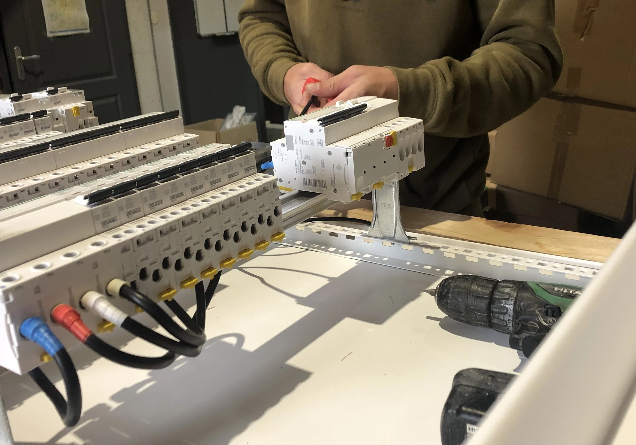 Une personne travaille sur un tableau électrique avec des composants haute tension, un tournevis à la main, dans un atelier.