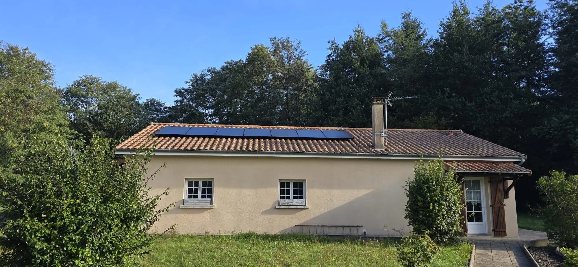 Maison avec toit en tuiles, panneaux solaires et cheminée, entourée d'arbres, sous un ciel bleu.