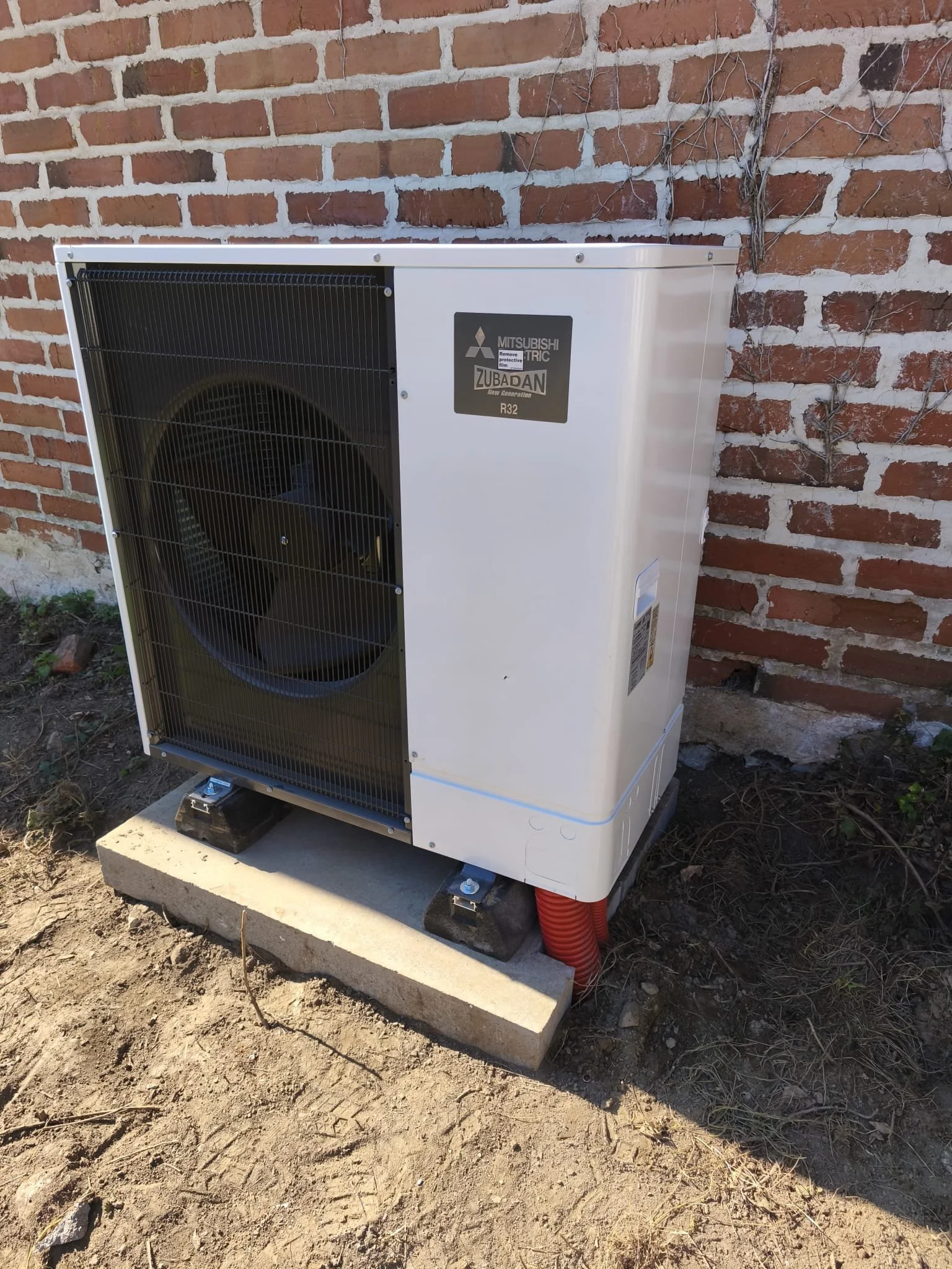 Un unité extérieure de climatisation Mitsubishi Zubadan R32 installée à l'extérieur d'un bâtiment, posée sur un support en béton, avec un mur en briques en arrière-plan.