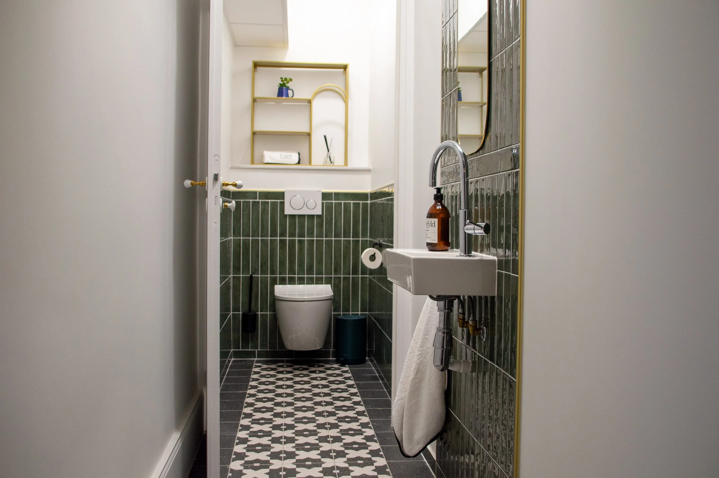 Petite salle de bain avec un mur en carrelage vert foncé, un lavabo blanc, un miroir rectangulaire, des étagères en bois avec des plantes et des objets décoratifs, un WC suspendu avec un bouton flush circulaire, un porte-rouleau de papier toilette, et un tapis au sol avec un motif noir et blanc.