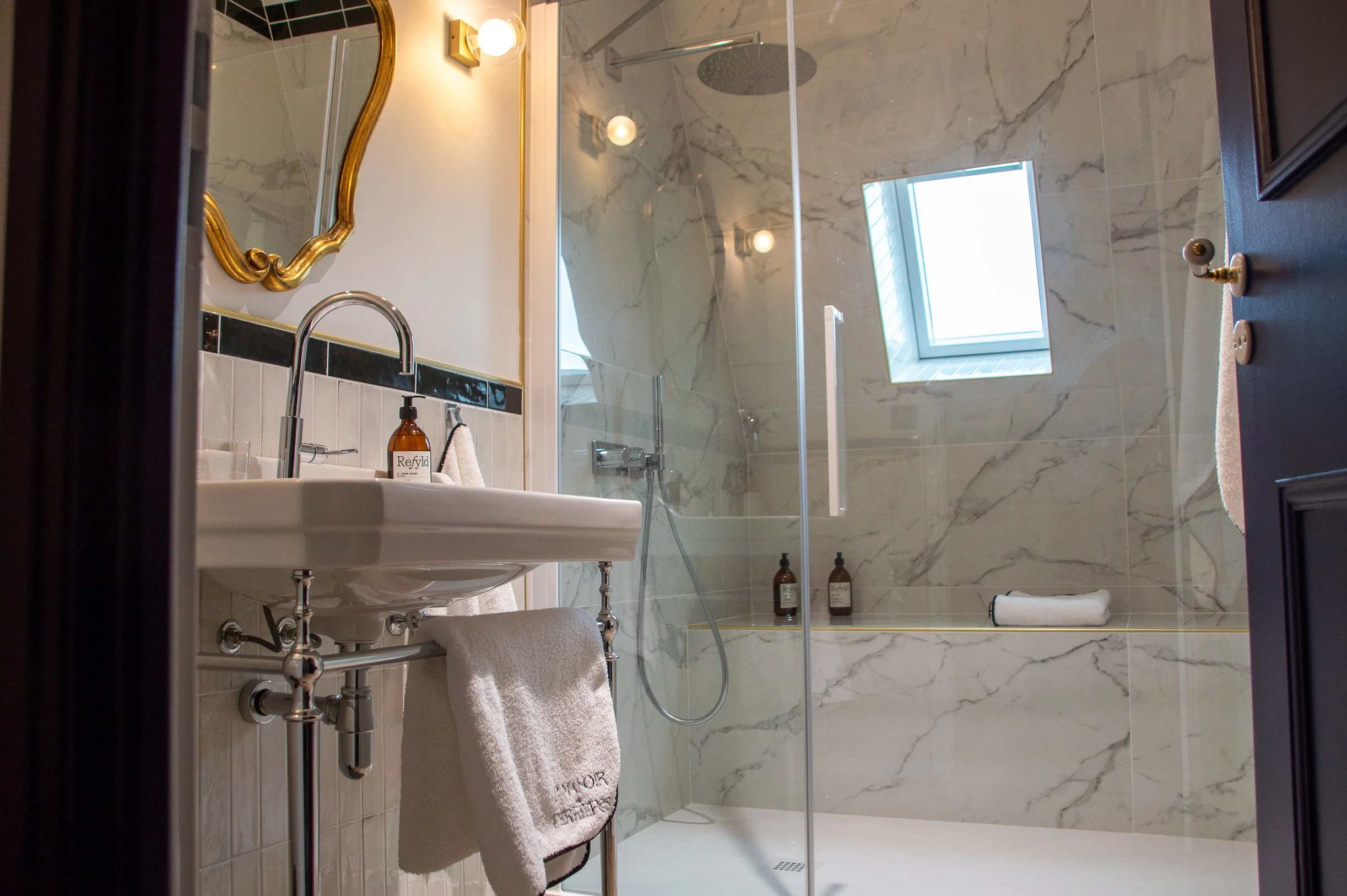 Salle de bain avec mur en marbre, douche à l'italienne avec porte en verre, fenêtre de toit, lavabo avec un miroir doré, serviette blanche suspendue à la barre sous le lavabo, produits de bain sur la paroi de la douche