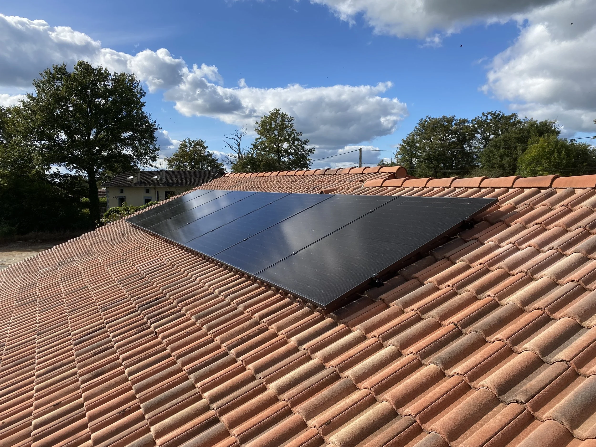 Panneaux solaires installés sur un toit en tuiles en terre cuite, avec un ciel bleu et des nuages dans le ciel, et des arbres en arrière-plan.