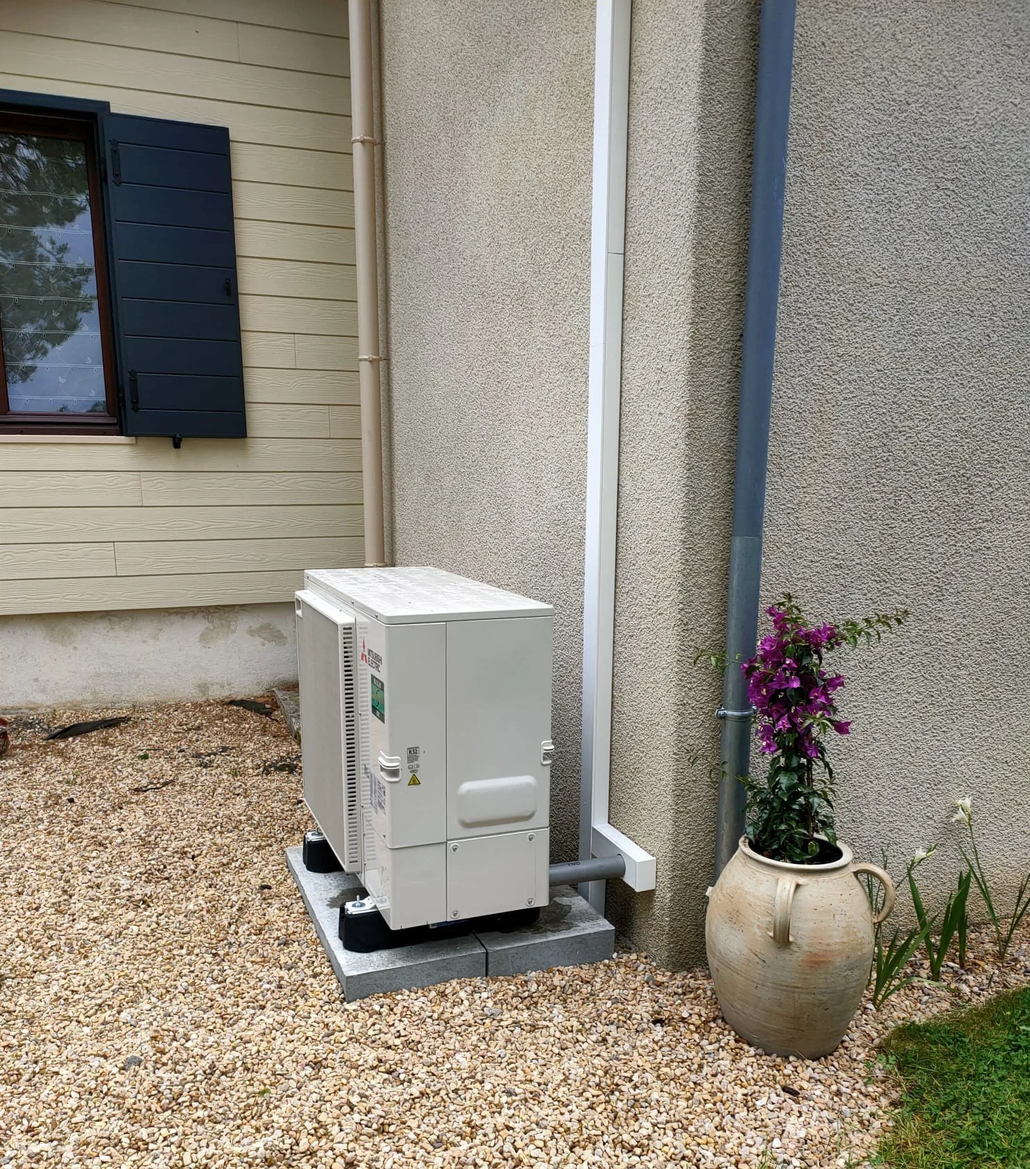 Chaudière extérieure située sur une plateforme en béton, à côté d'une plante en pot avec des fleurs violettes, contre un mur beige d'une maison avec une fenêtre à volets bleus.