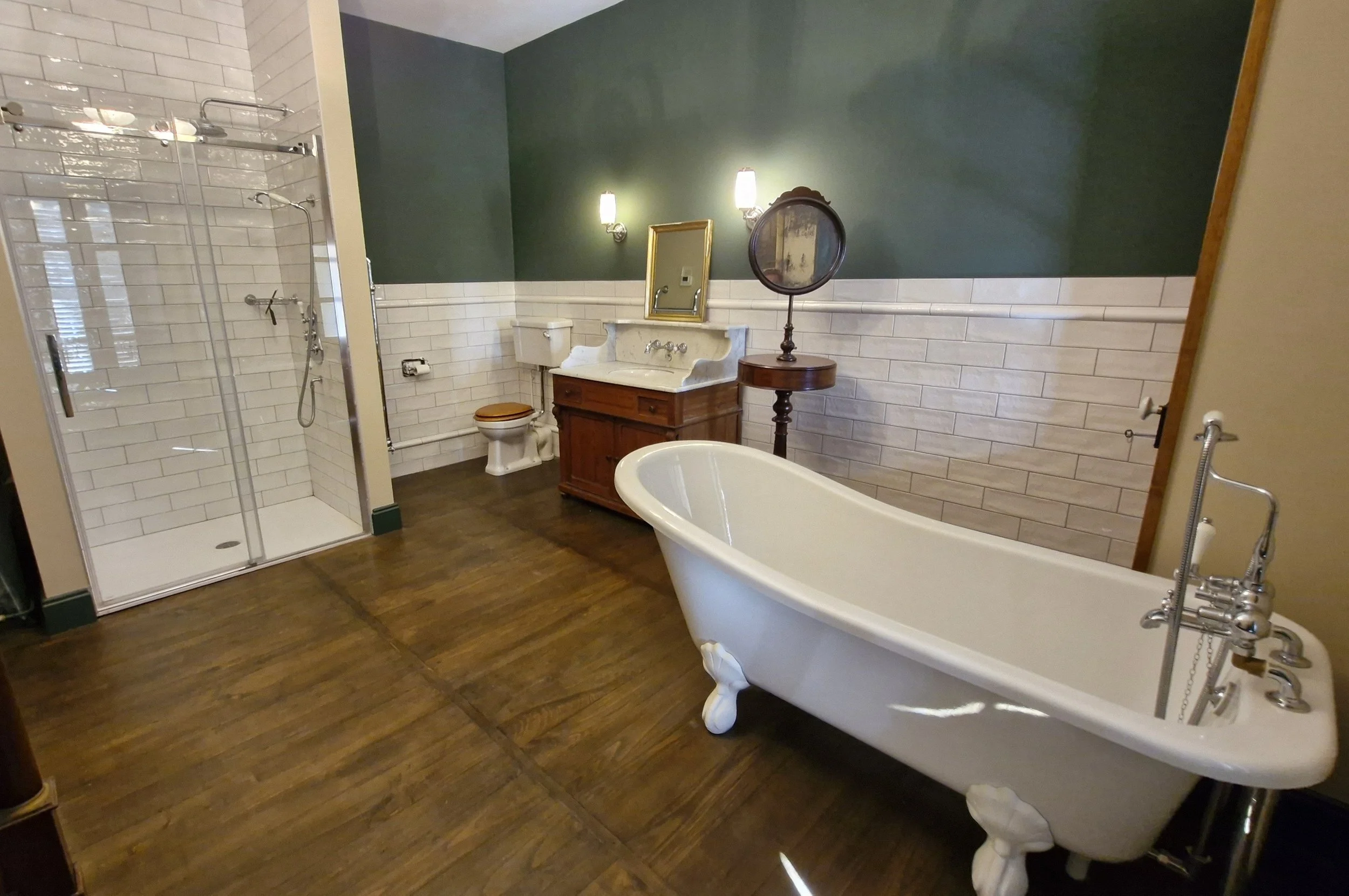 Salle de bain avec baignoire, cabine de douche, toilette et meuble avec lavabo, murs peints en vert et blanc, plancher en bois.