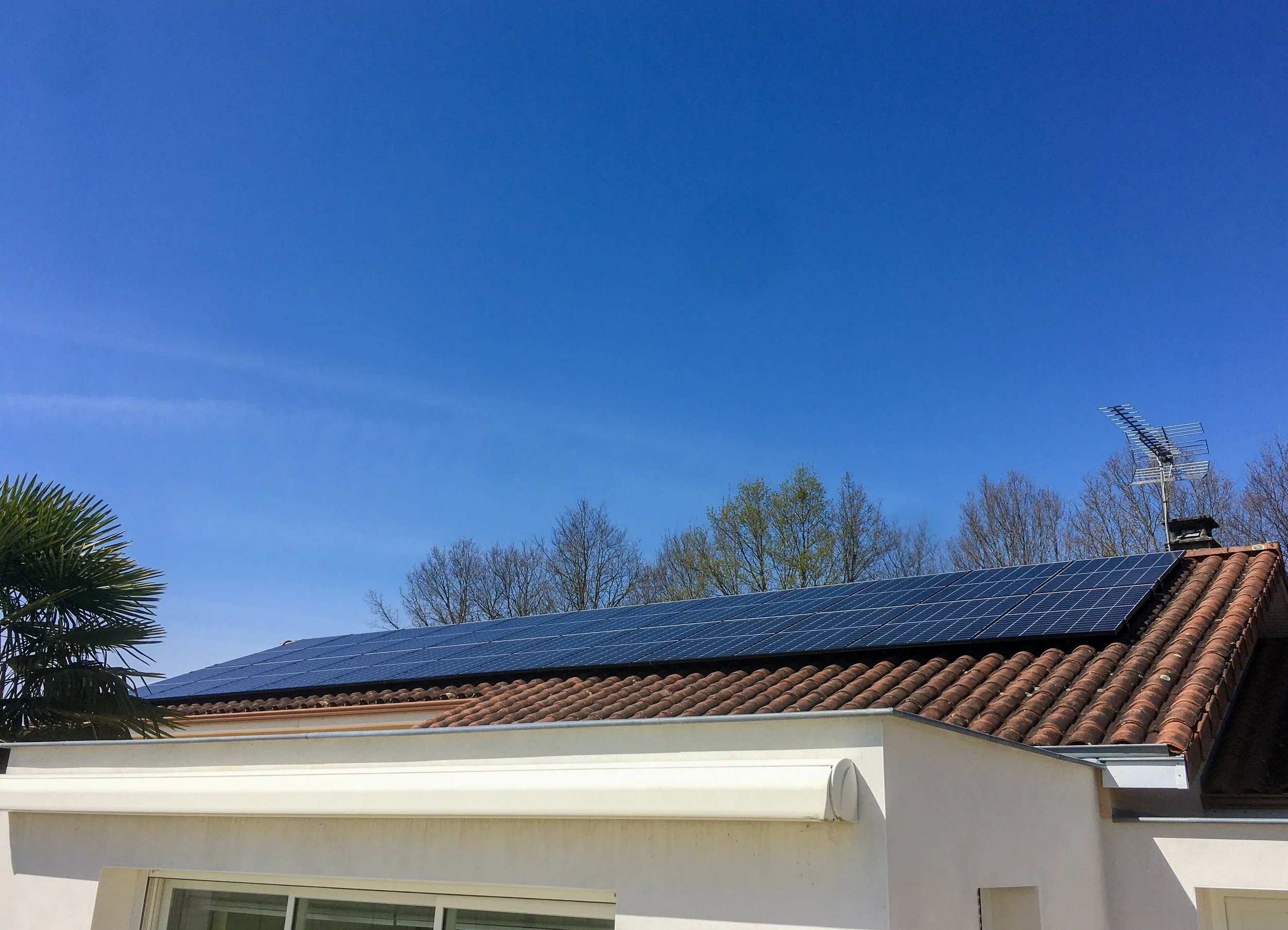 Toit de maison avec panneaux solaires, tuiles en terre cuite, antenne TV et ciel bleu.