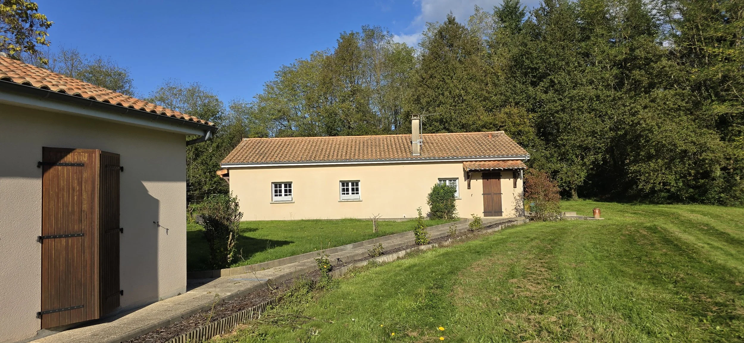 Une maison avec un toit en tuiles rouges, des fenêtres blanches, entourée d'arbres et d'herbe verte, sous un ciel bleu clair.