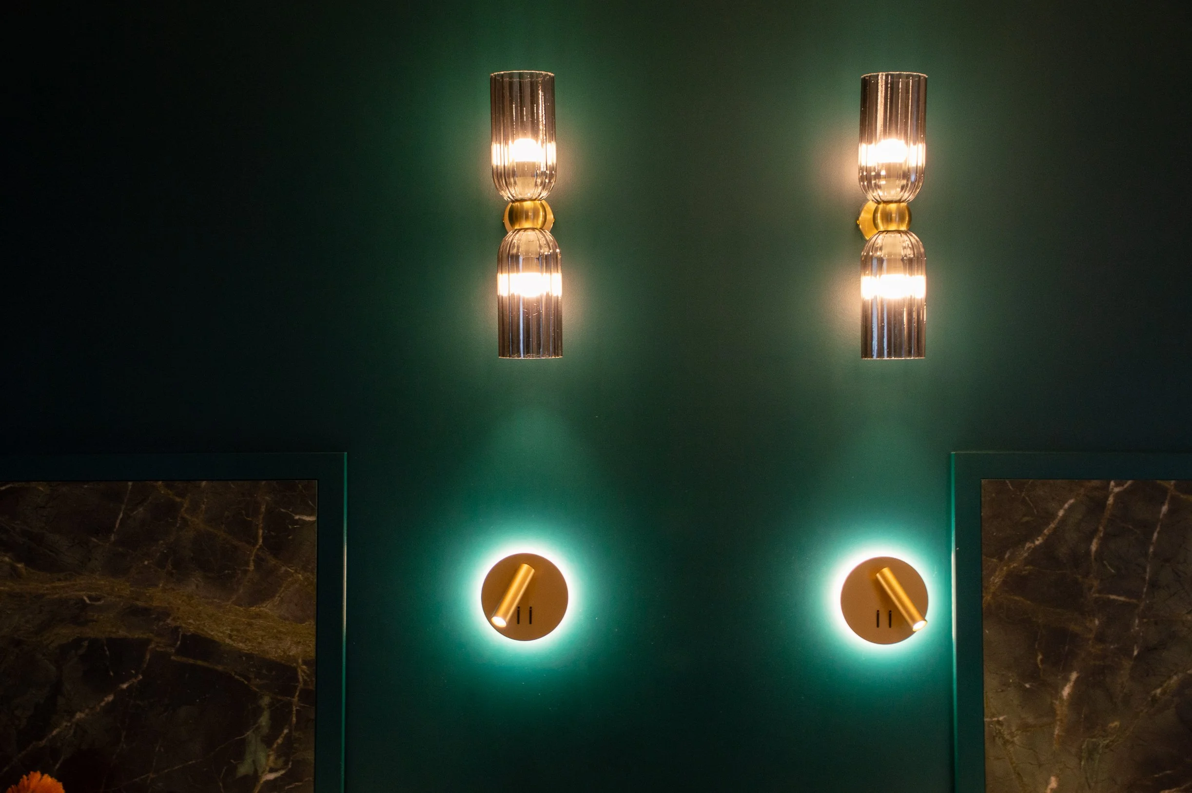 Deux appliques murales modernes en métal argenté avec lumière chaude, fixées à un mur vert foncé, avec deux luminaires ronds en bas émettant une lumière douce