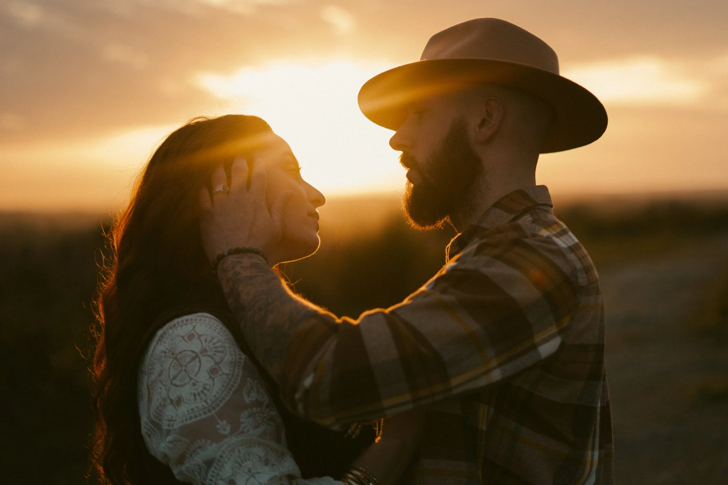 Un homme et une femme regardent intensément l'un l'autre lors d'un coucher de soleil, l'homme porte un chapeau et une chemise à carreaux, la femme a les cheveux longs et chatoyants, en train de se tenir face à face dans un paysage naturel.