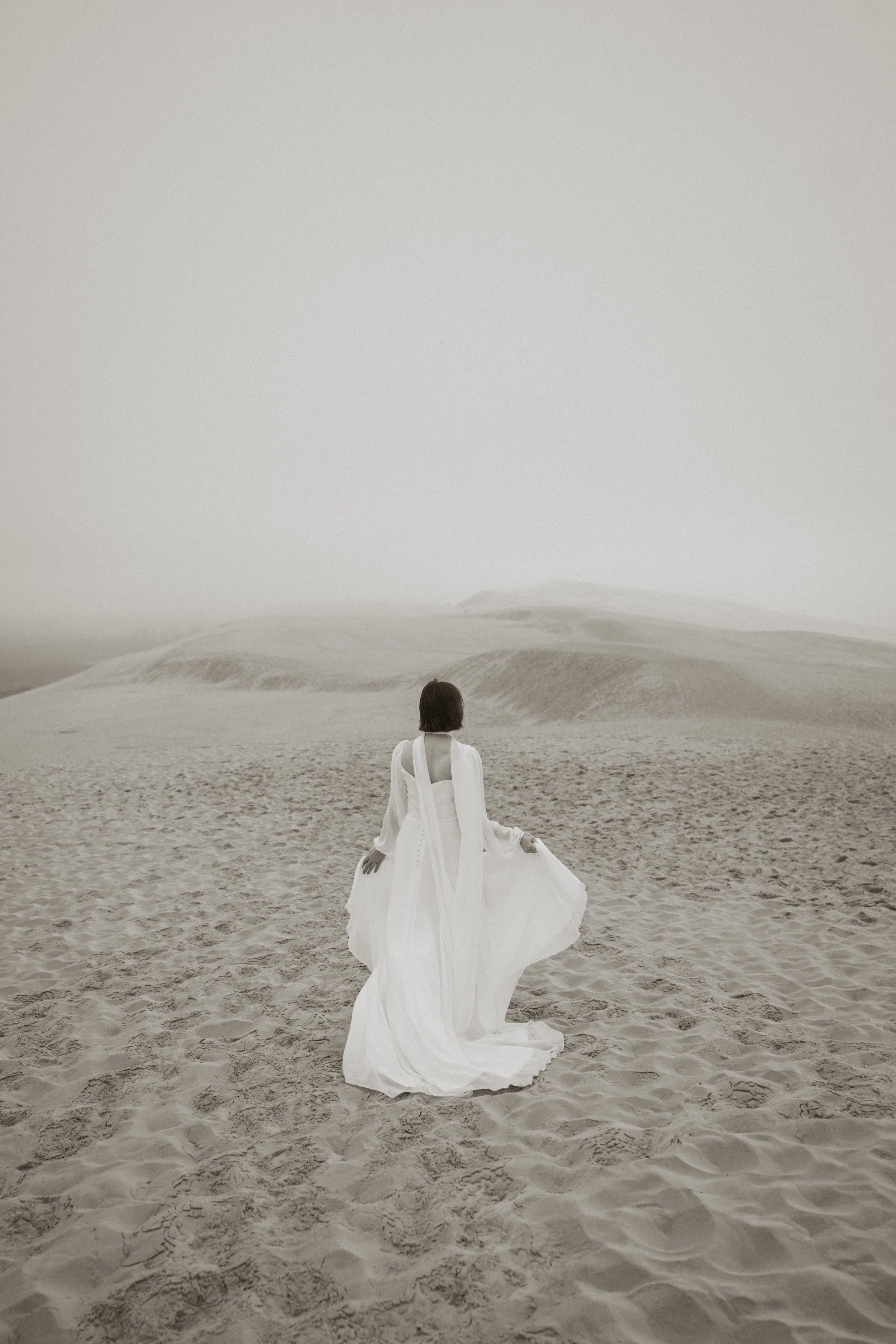 Une femme en robe blanche dans un désert, vue de dos, sous un ciel gris et nuageux.