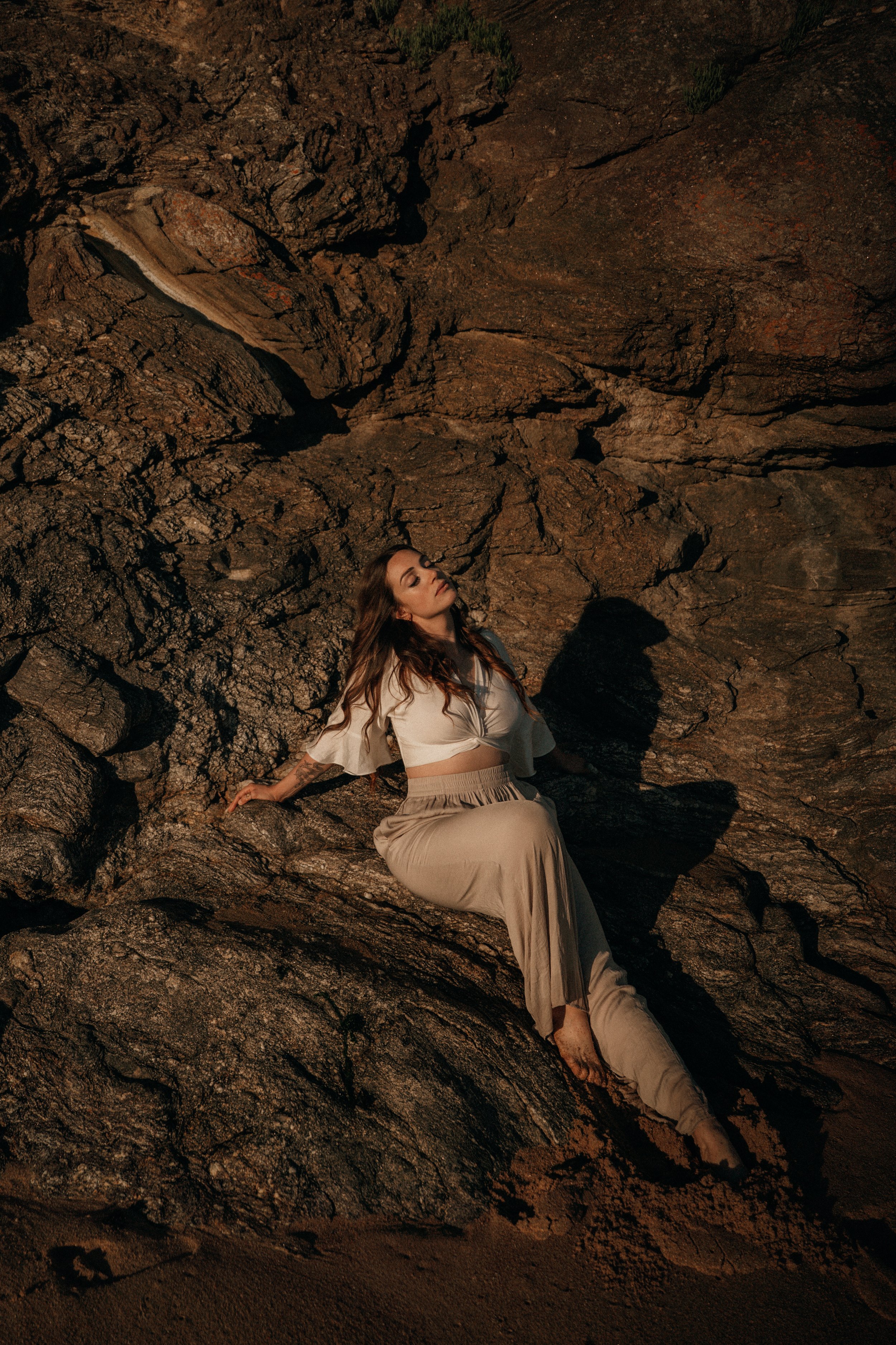 Femme assise contre des rochers au coucher du soleil. Vêtements beige, cheveux roux, regard détaché.