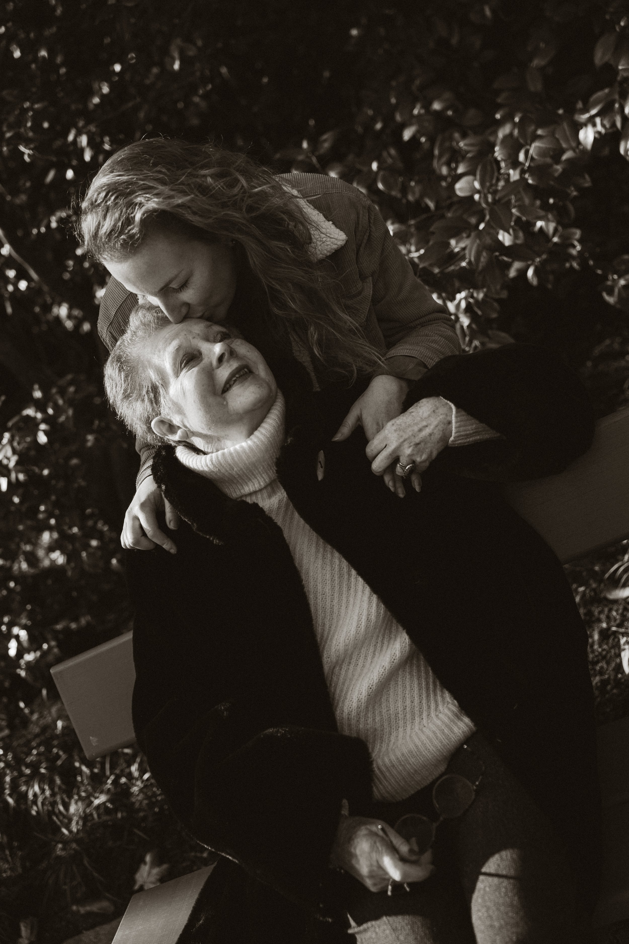 Une jeune femme embrasse une vieille femme souriante, toutes deux assises parmi des feuilles d'arbres, en noir et blanc.