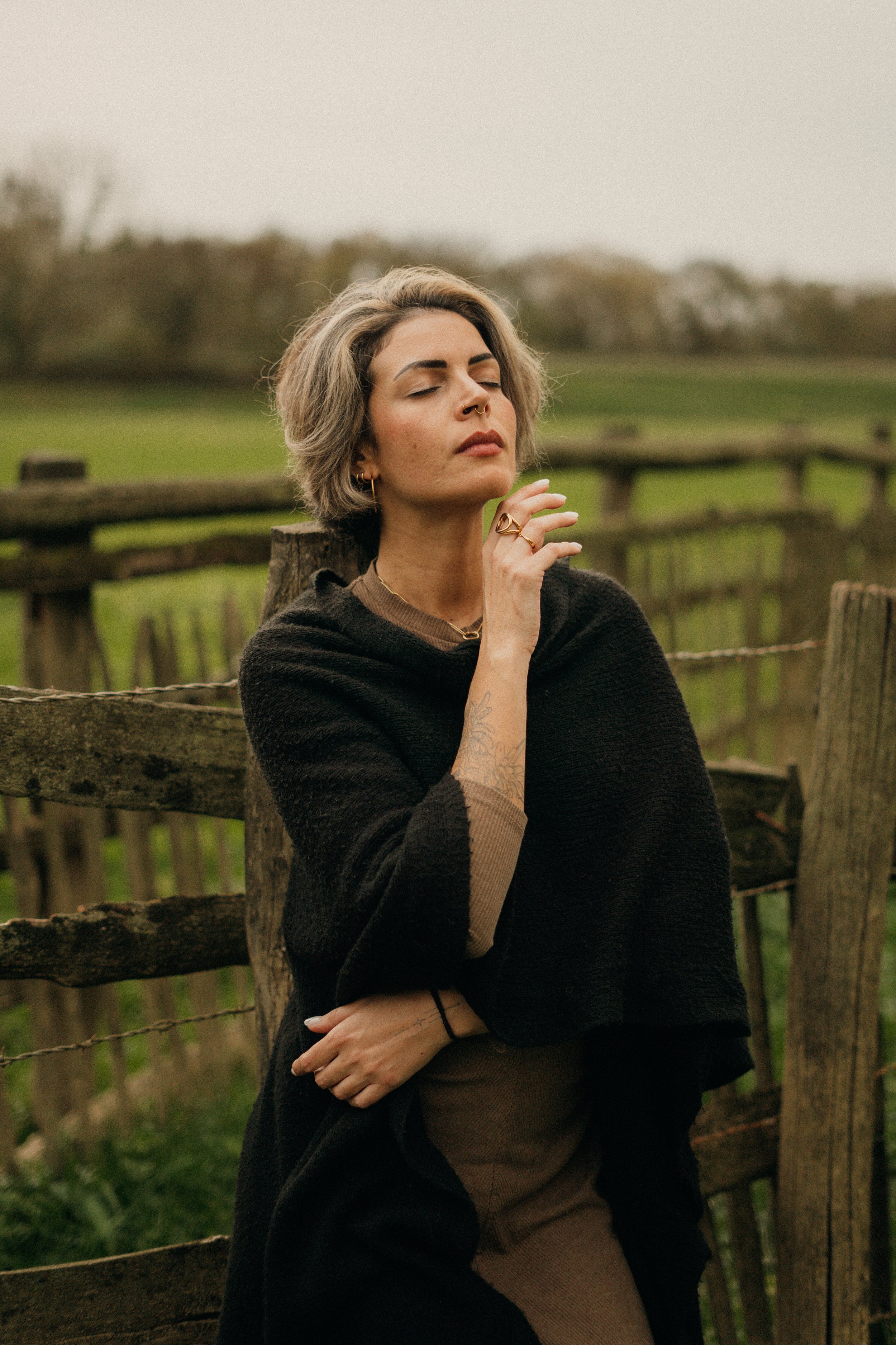 Une femme aux cheveux gris court, portant des vêtements beige et noir, avec des tatouages, est debout près d'une clôture en bois dans un champ vert, avec un ciel gris en arrière-plan.