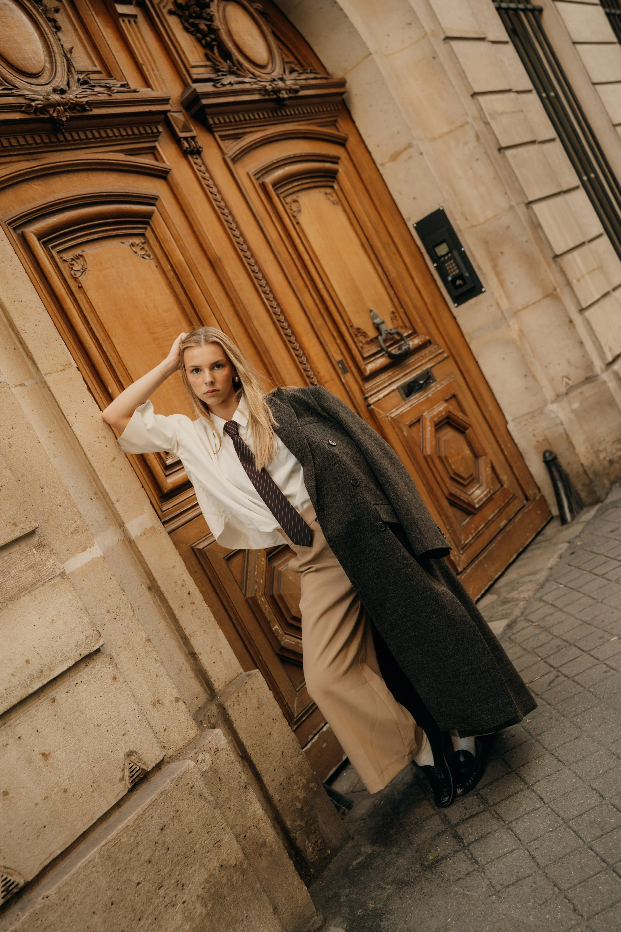 Jeune femme posant devant une grande porte en bois ornée de détails sculptés, dans un style architectural classique.