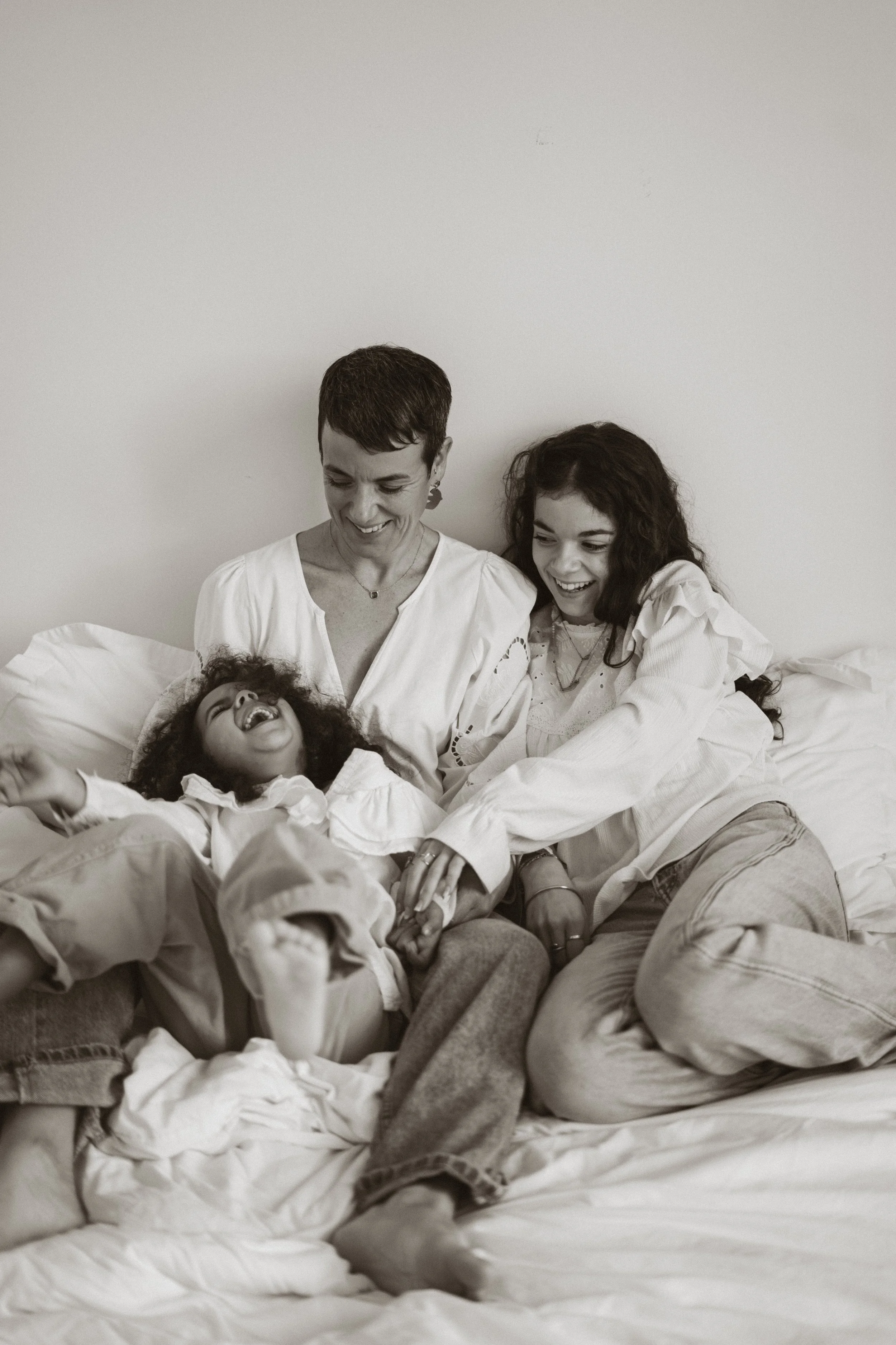 Une famille souriante et heureuse, composée de deux femmes et d'une jeune fille, assise sur un lit. La scène est en noir et blanc.