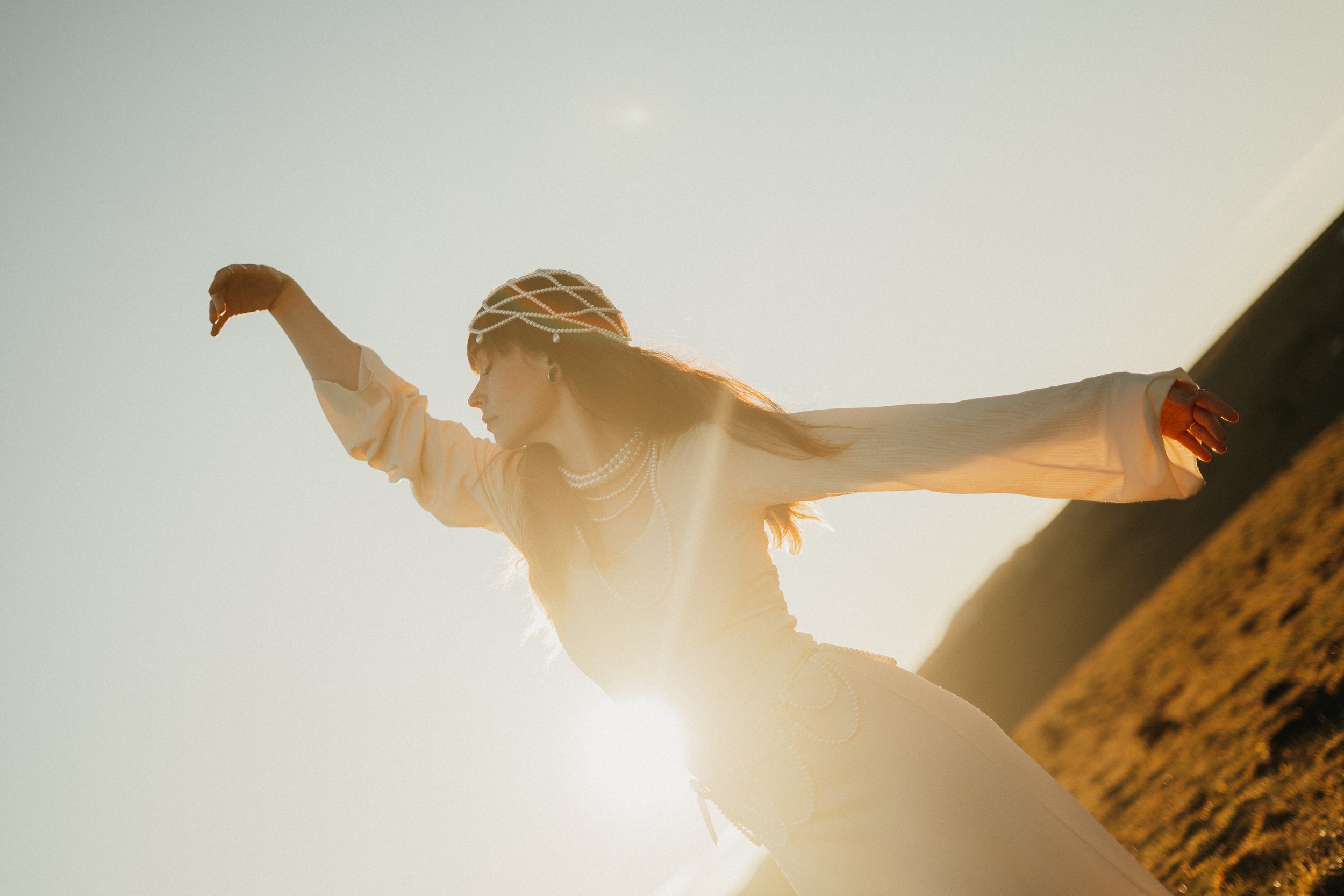 Une femme portant une robe blanche et des bijoux, debout dans un paysage en plein air au soleil couchant.