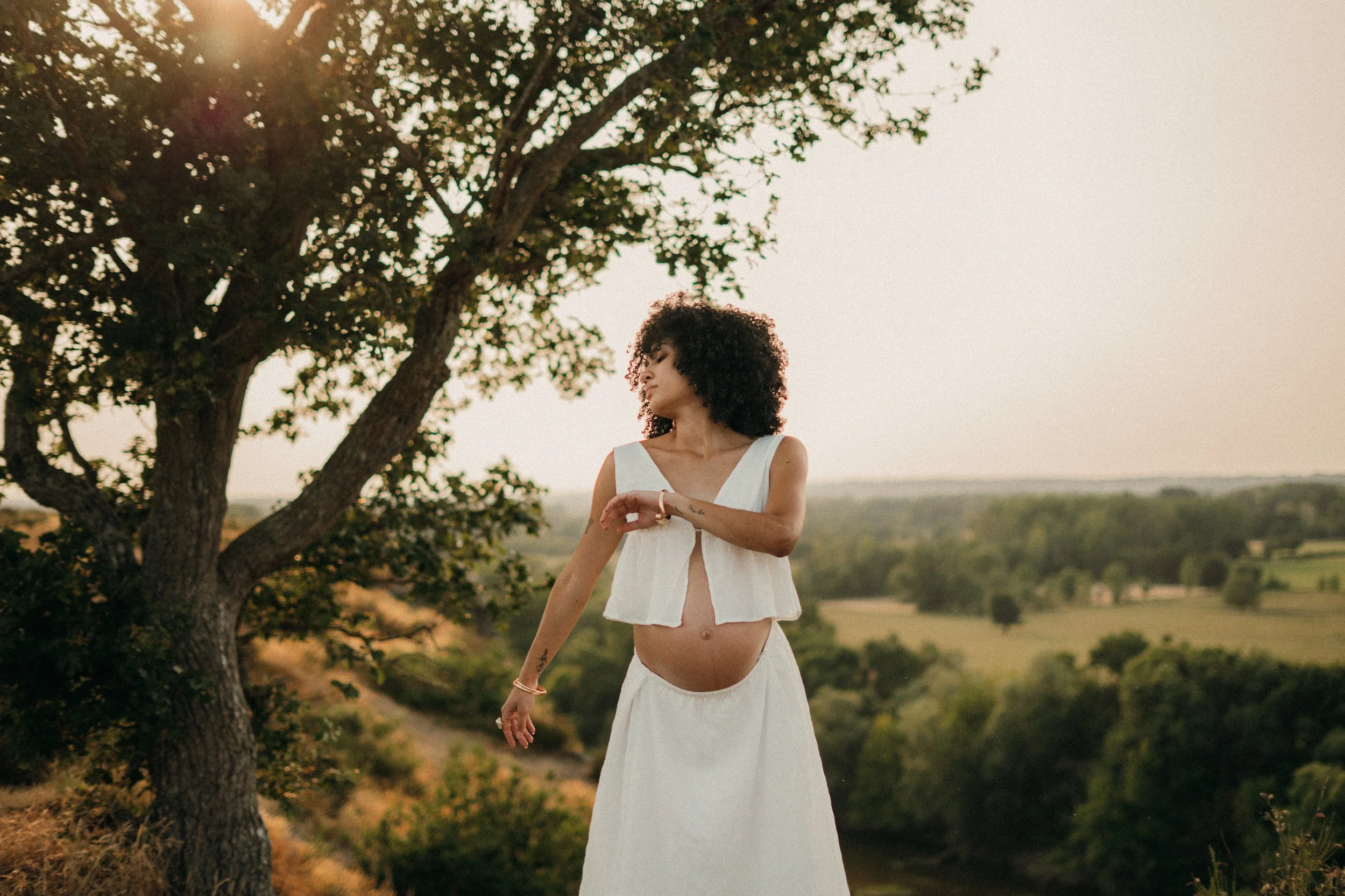 Femme enceinte portant une robe blanche et un haut blanc léger, posant à côté d'un arbre lors d'une séance photo en plein air au coucher du soleil, avec un paysage de champs et d'arbres en arrière-plan.