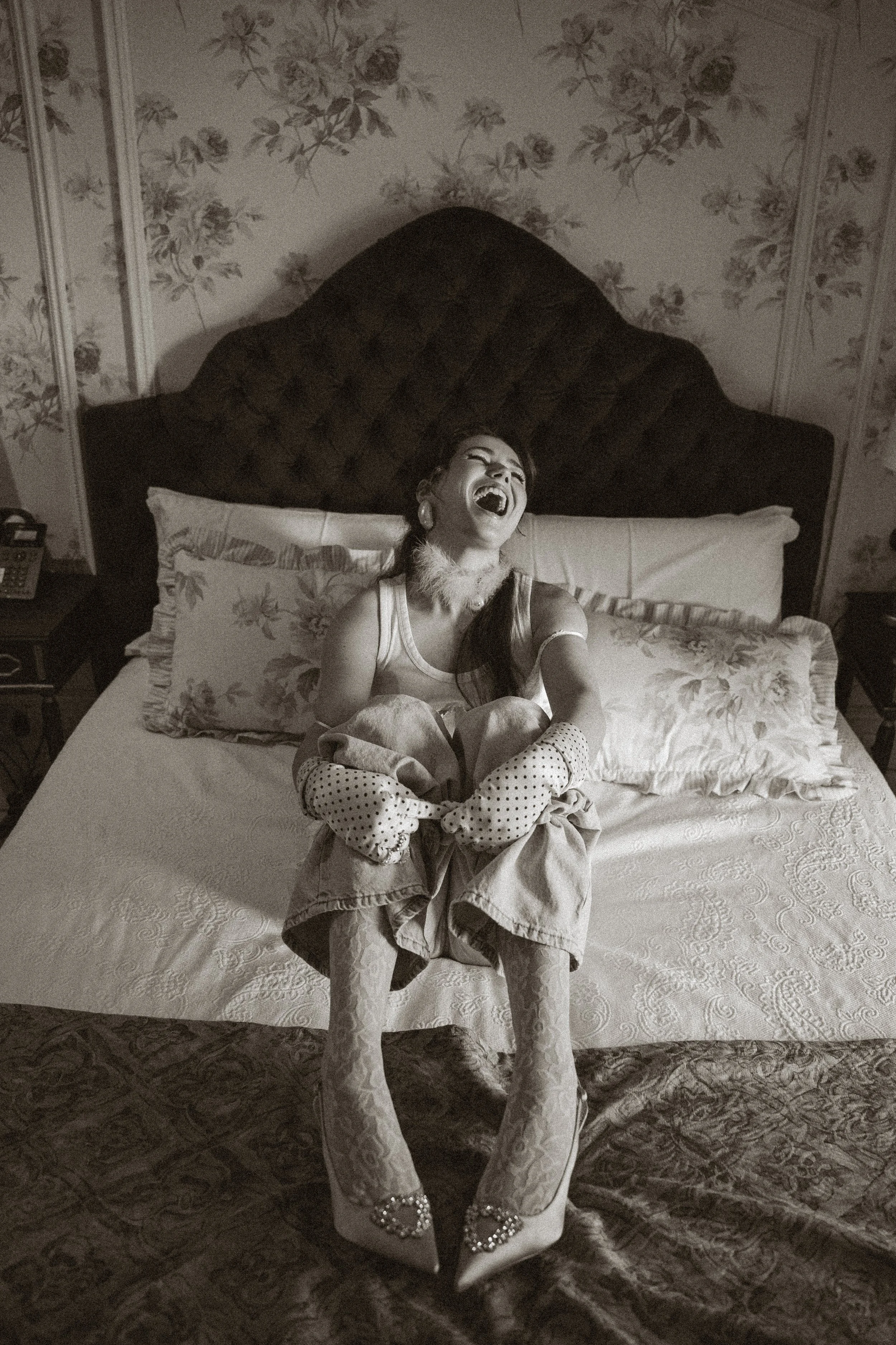 Une femme riant assise sur un lit dans une chambre décorée de papier peint floral.