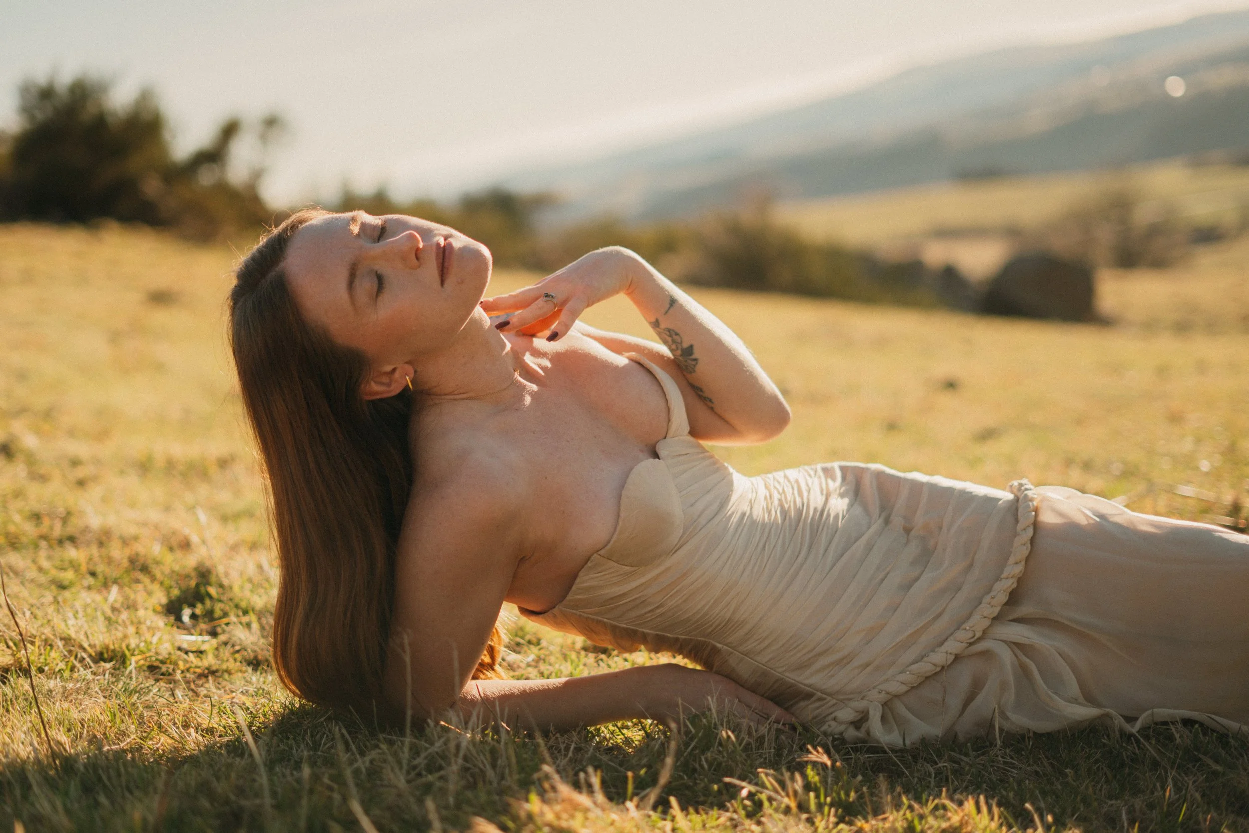 Femme allongée dans un champ, profitant du soleil, avec un paysage de montagnes en arrière-plan.