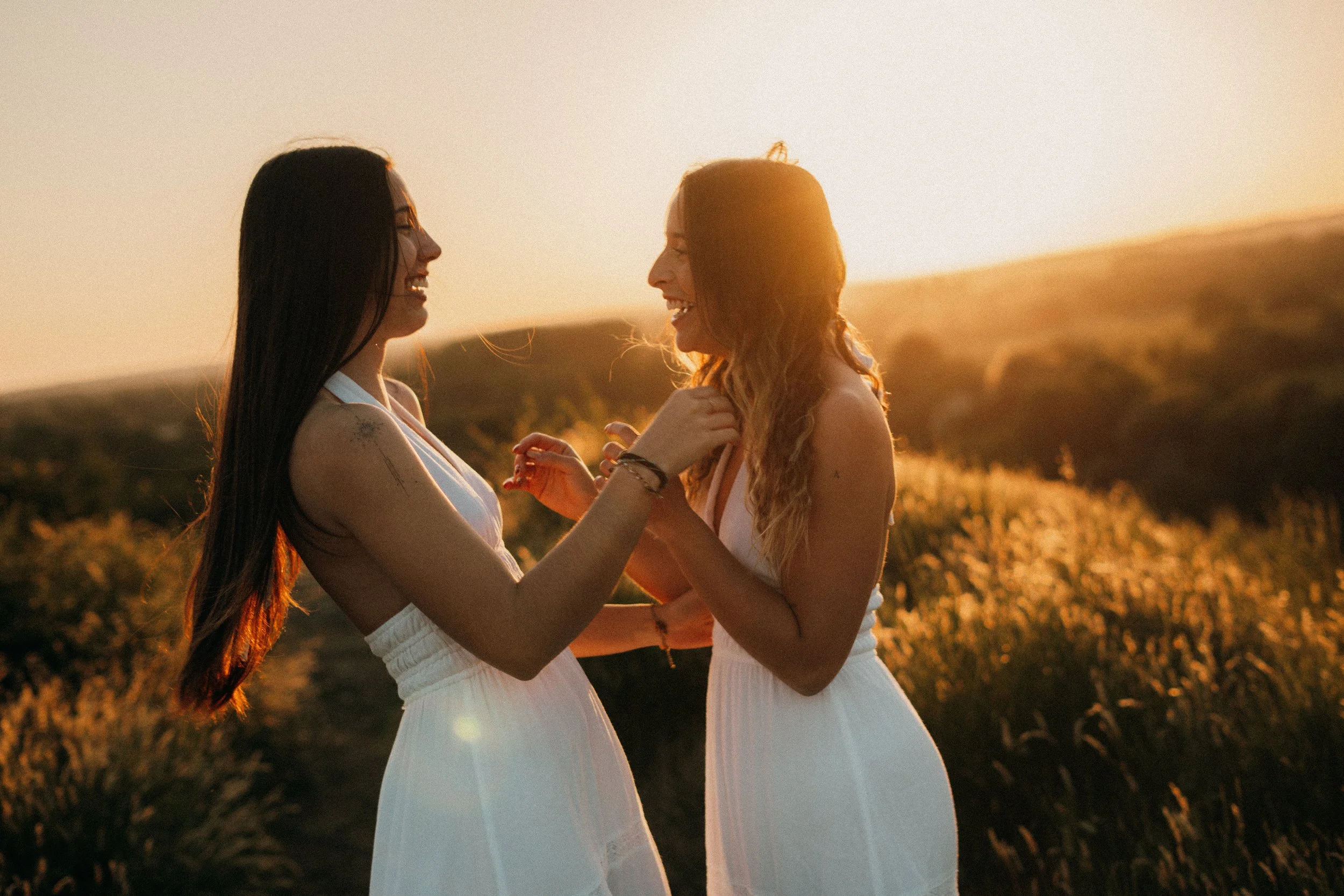 Deux femmes souriantes en robes blanches se tiennent dans un champ lors d'un coucher de soleil, échangeant un moment joyeux.