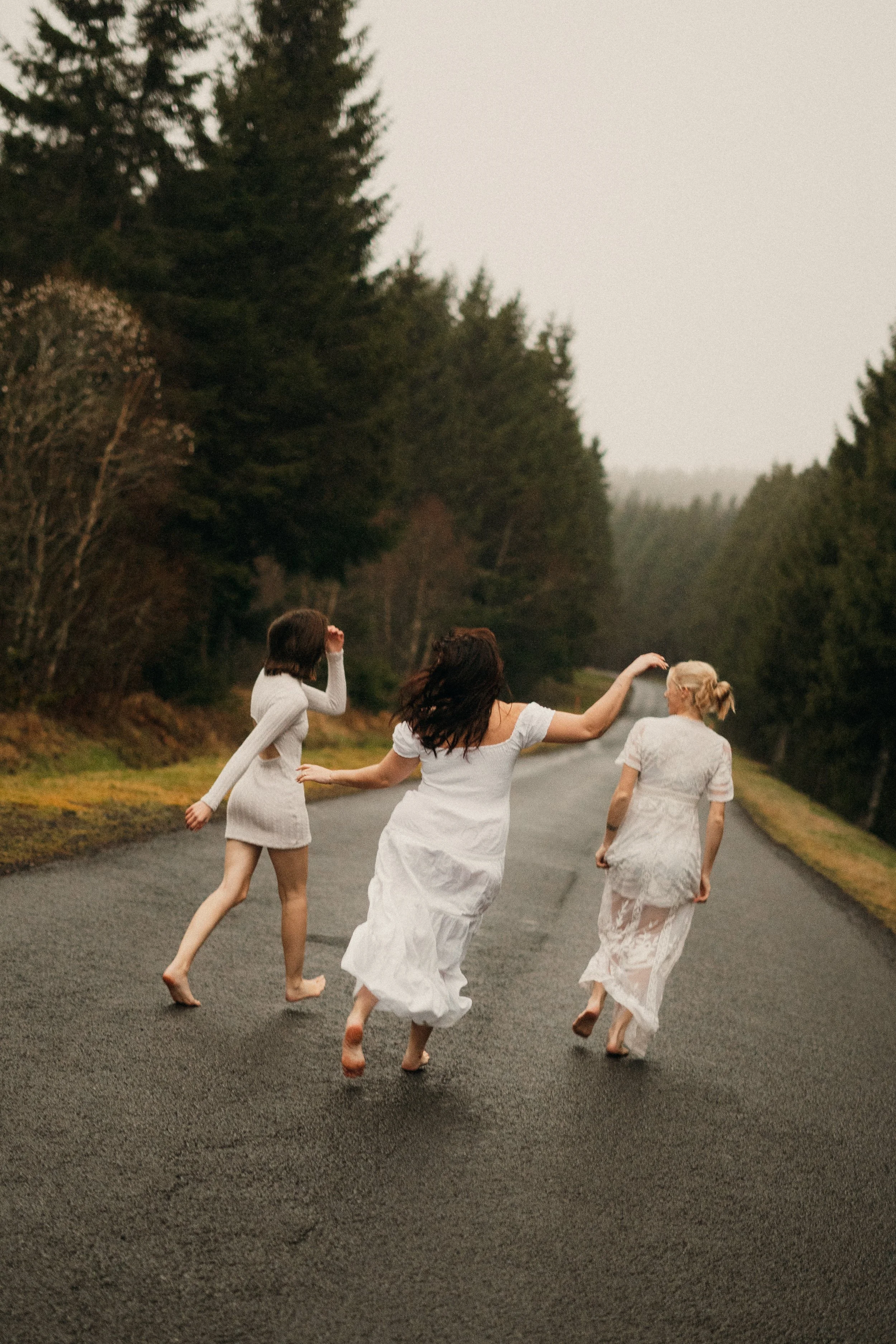 Trois femmes portant des robes blanches marchent ou courent pieds nus sur une route entourée d'arbres, dans une ambiance naturelle et paisible.