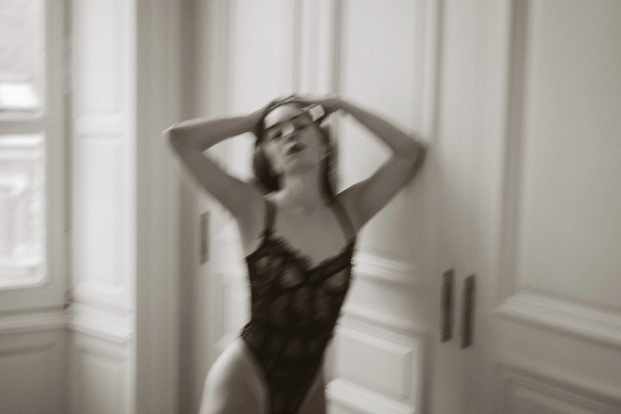 Une femme portant un bodysuit en dentelle noire, légèrement floue, dans une pièce lumineuse avec des murs blancs et une porte en arrière-plan.
