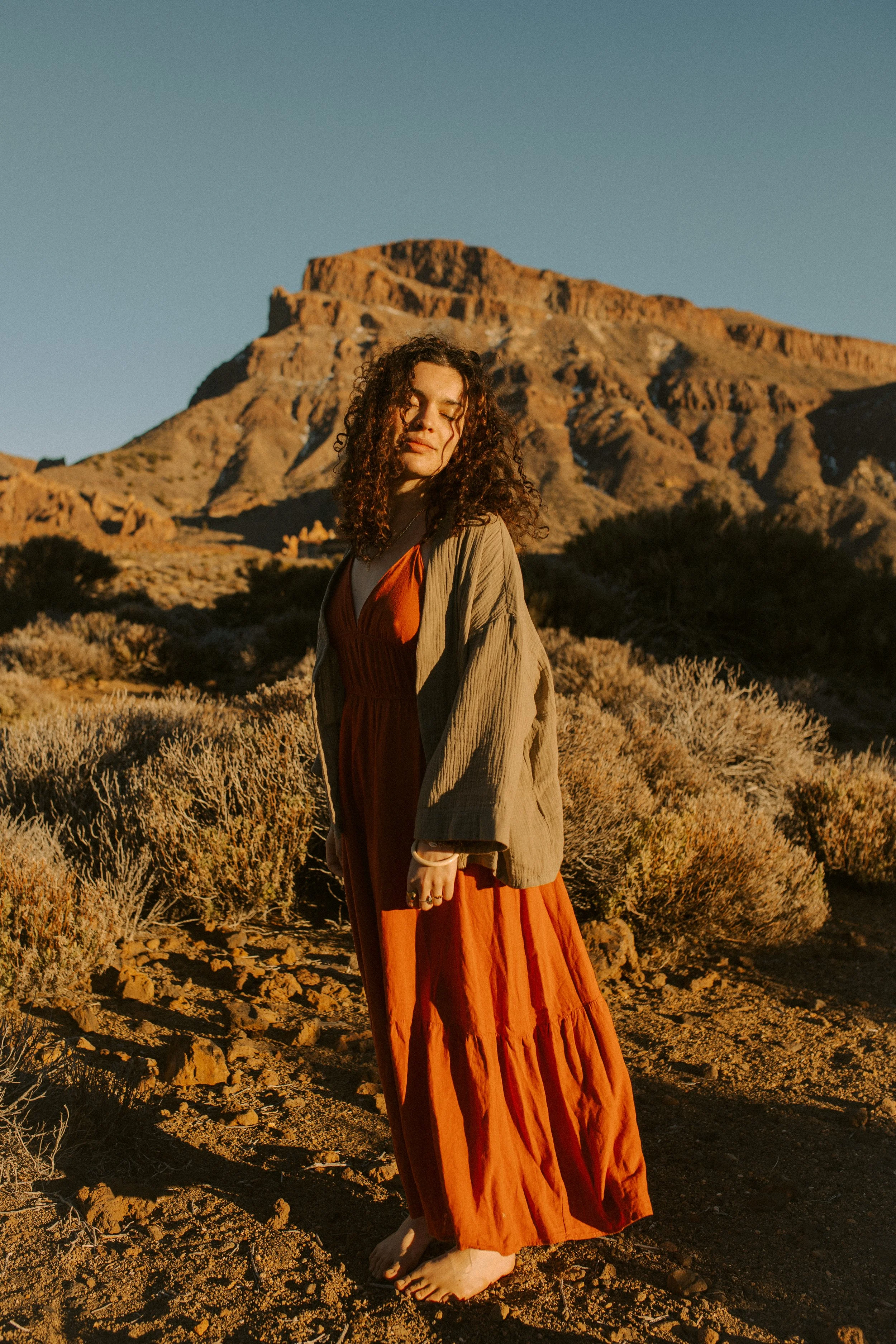 Jeune femme aux cheveux frisés en robe orange, portant une veste beige, debout dans un désert avec un rocher en arrière-plan au coucher du soleil.
