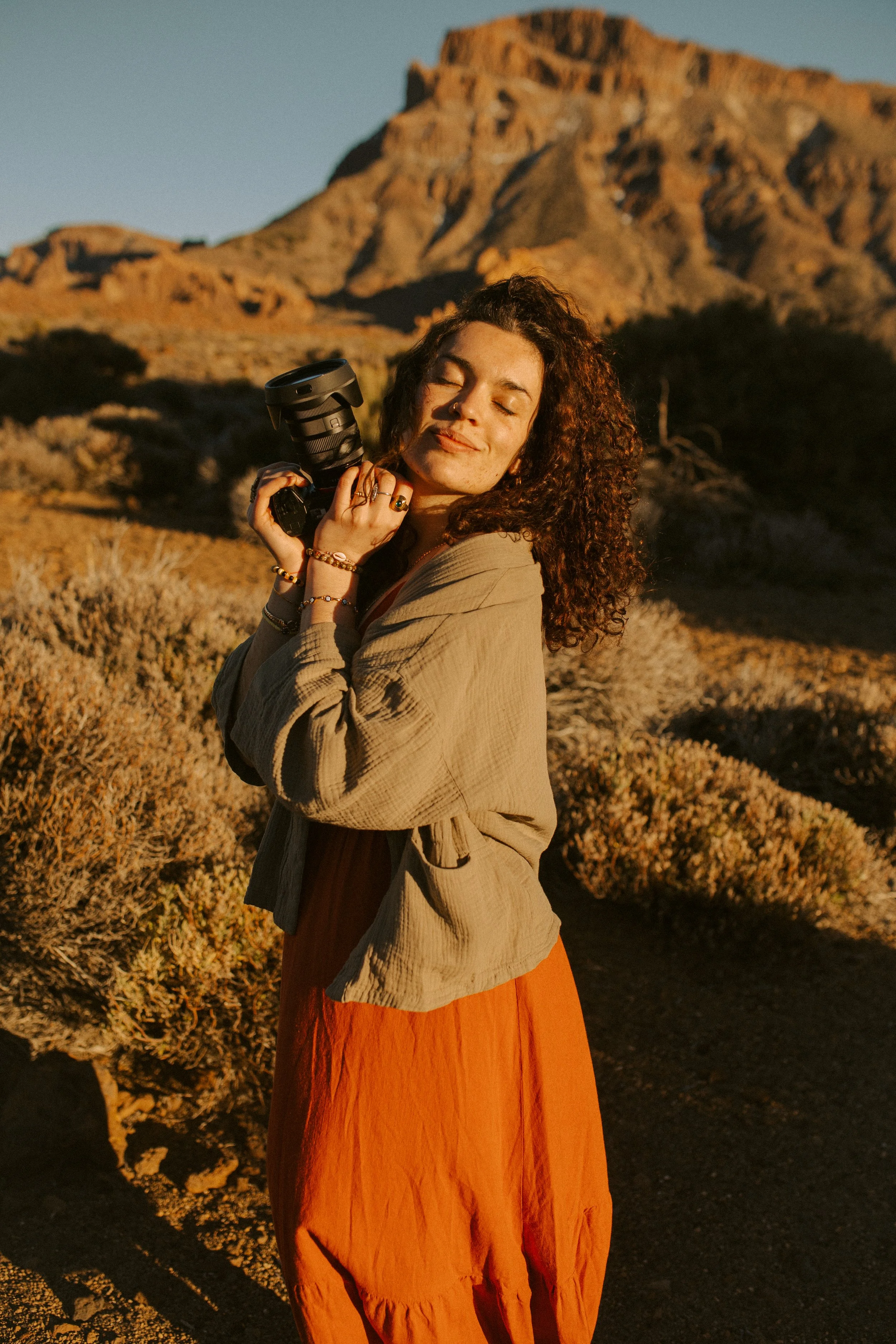 Une femme souriante avec des cheveux bouclés, tenant un appareil photo, dans un paysage désertique au coucher de soleil.