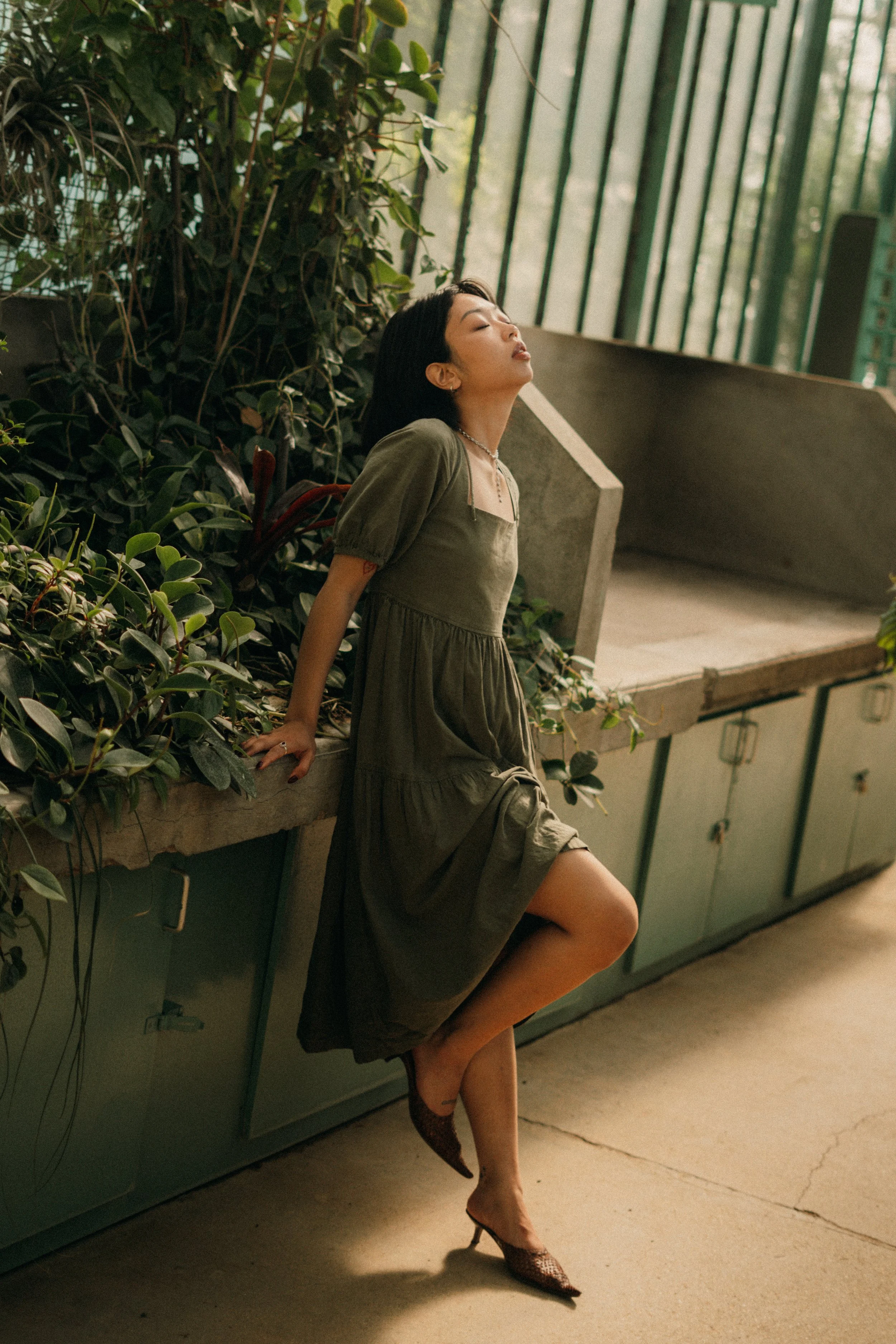 Une femme en robe verte s'appuie sur un comptoir en béton avec des plantes vertes derrière elle et des fenêtres vertes en arrière-plan, dans un environnement lumineux et calme.