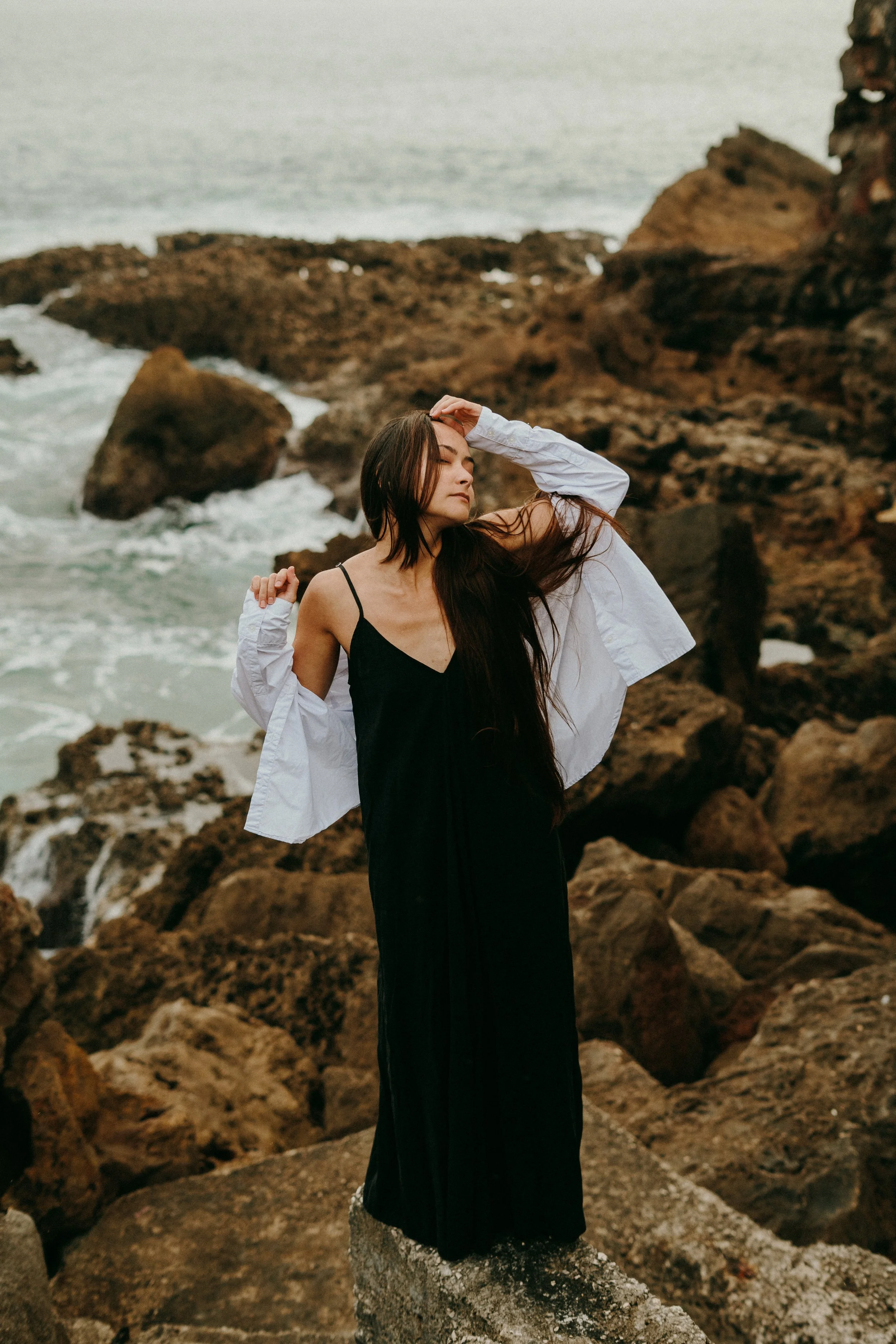 Jeune femme en robe noire et blazer blanc, posant sur des rochers près de la mer.