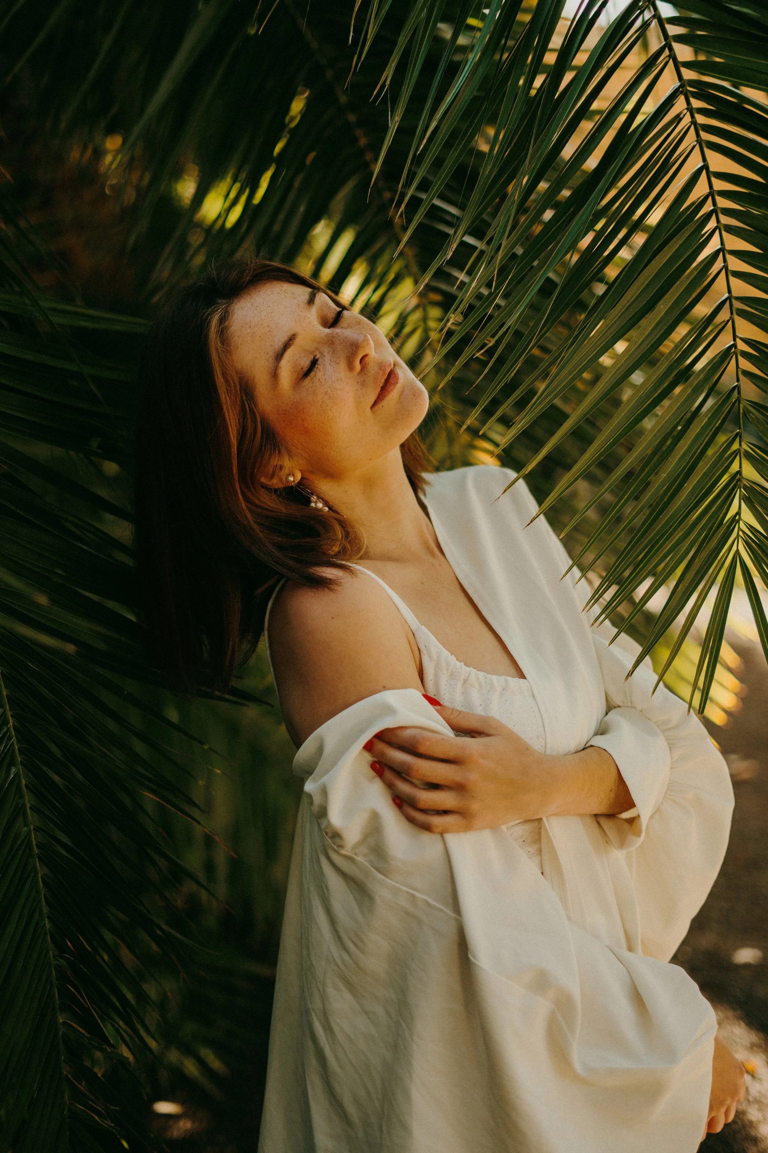 Une femme avec des cheveux bruns et des taches de rousseur, portant une robe blanche, sous des feuilles de palmier, yeux fermés et expression de relaxation.