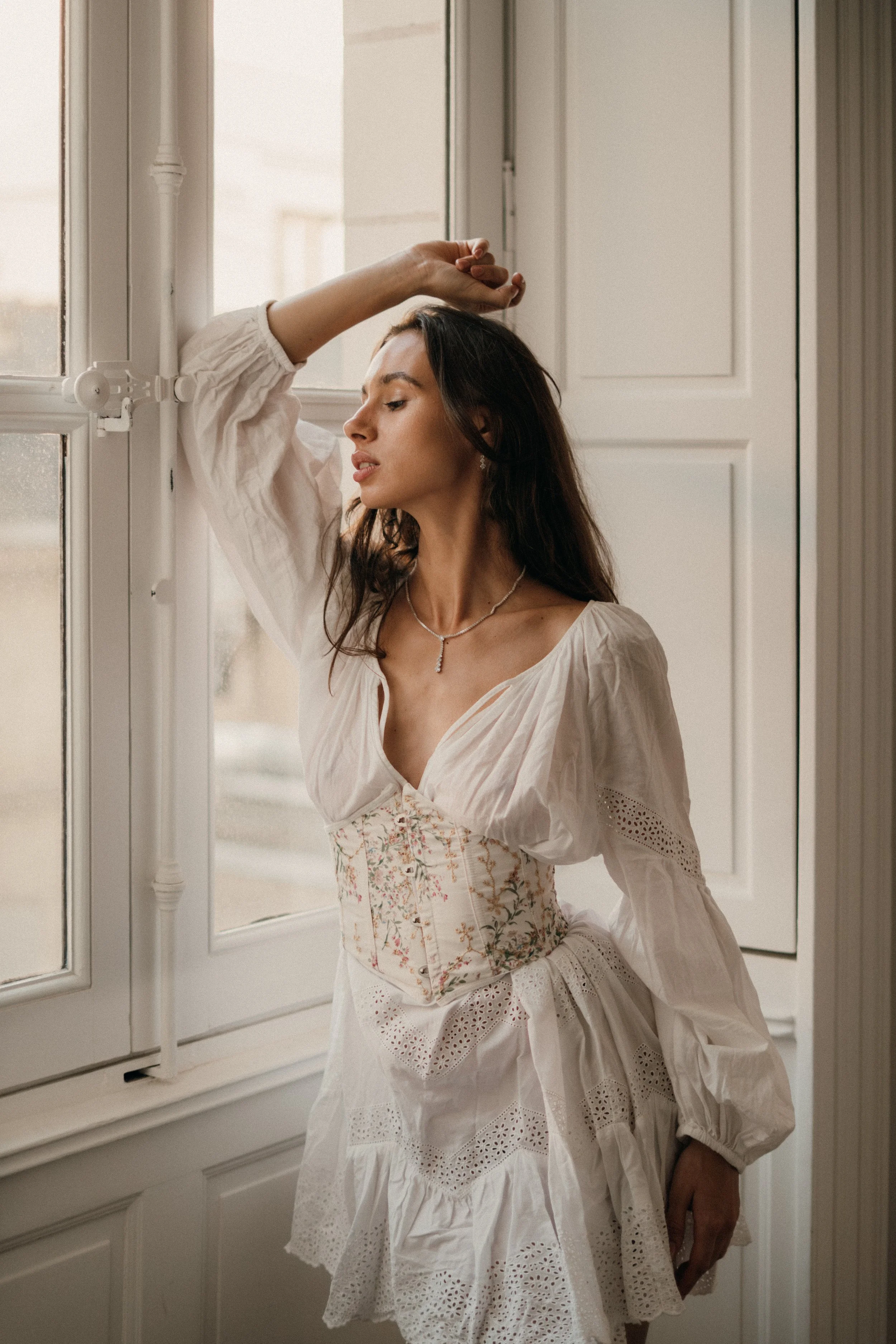 Femme en robe blanche avec corset floral, posant près d'une fenêtre en intérieur, dans une lumière douce.