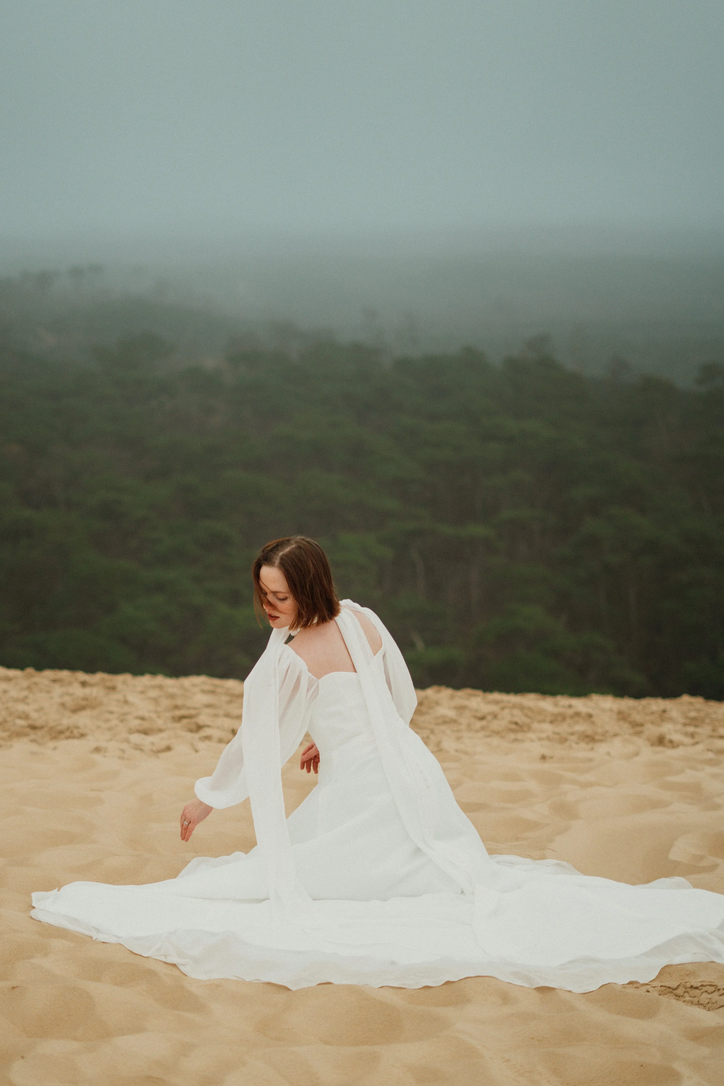 Une femme en robe blanche assise sur le sable dans un environnement naturel avec une forêt et un ciel nuageux en arrière-plan.