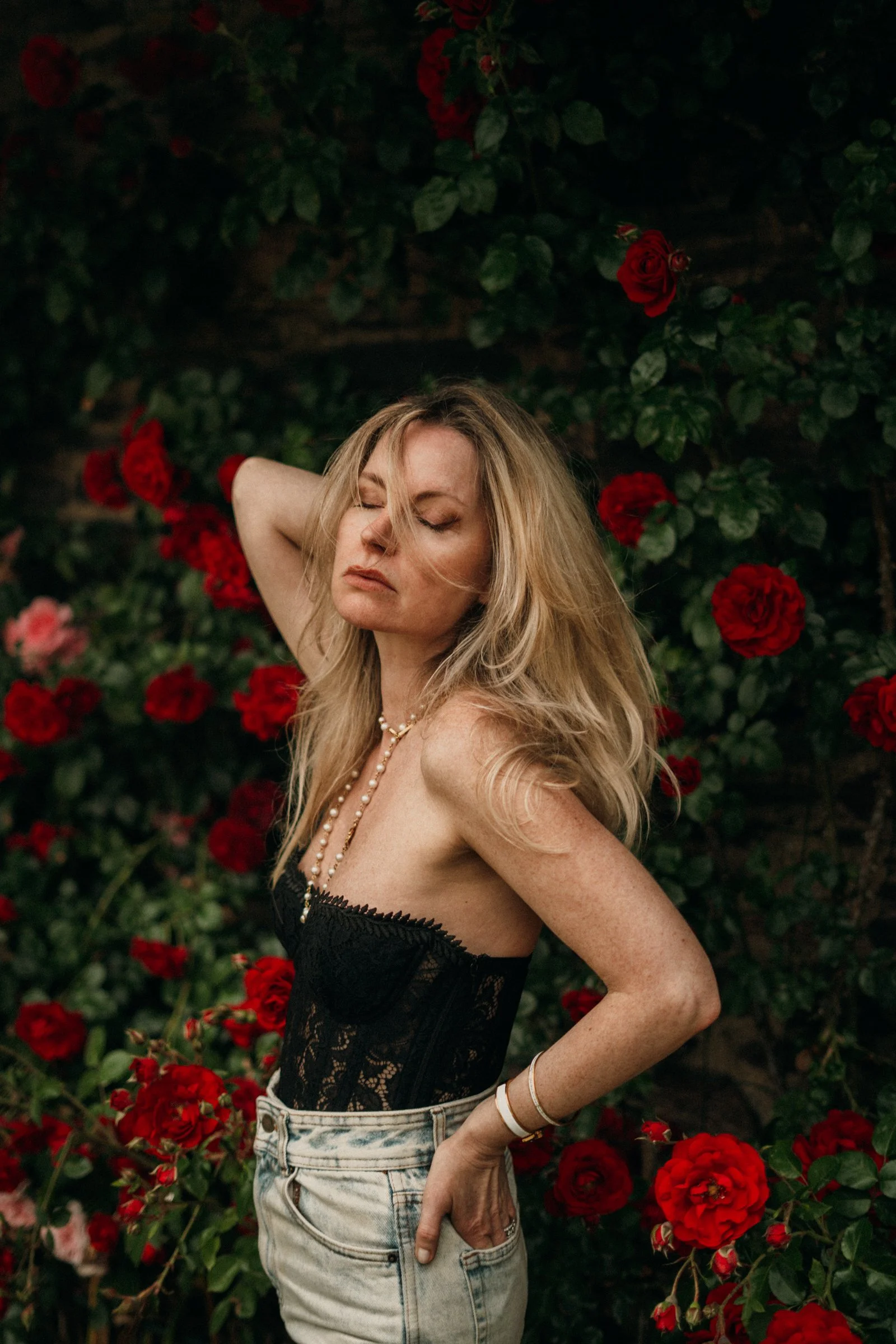 Femme avec longs cheveux blonds, portant un corset en dentelle noir, un jean délavé haut, et des bijoux, posant devant un mur de roses rouges dans un jardin