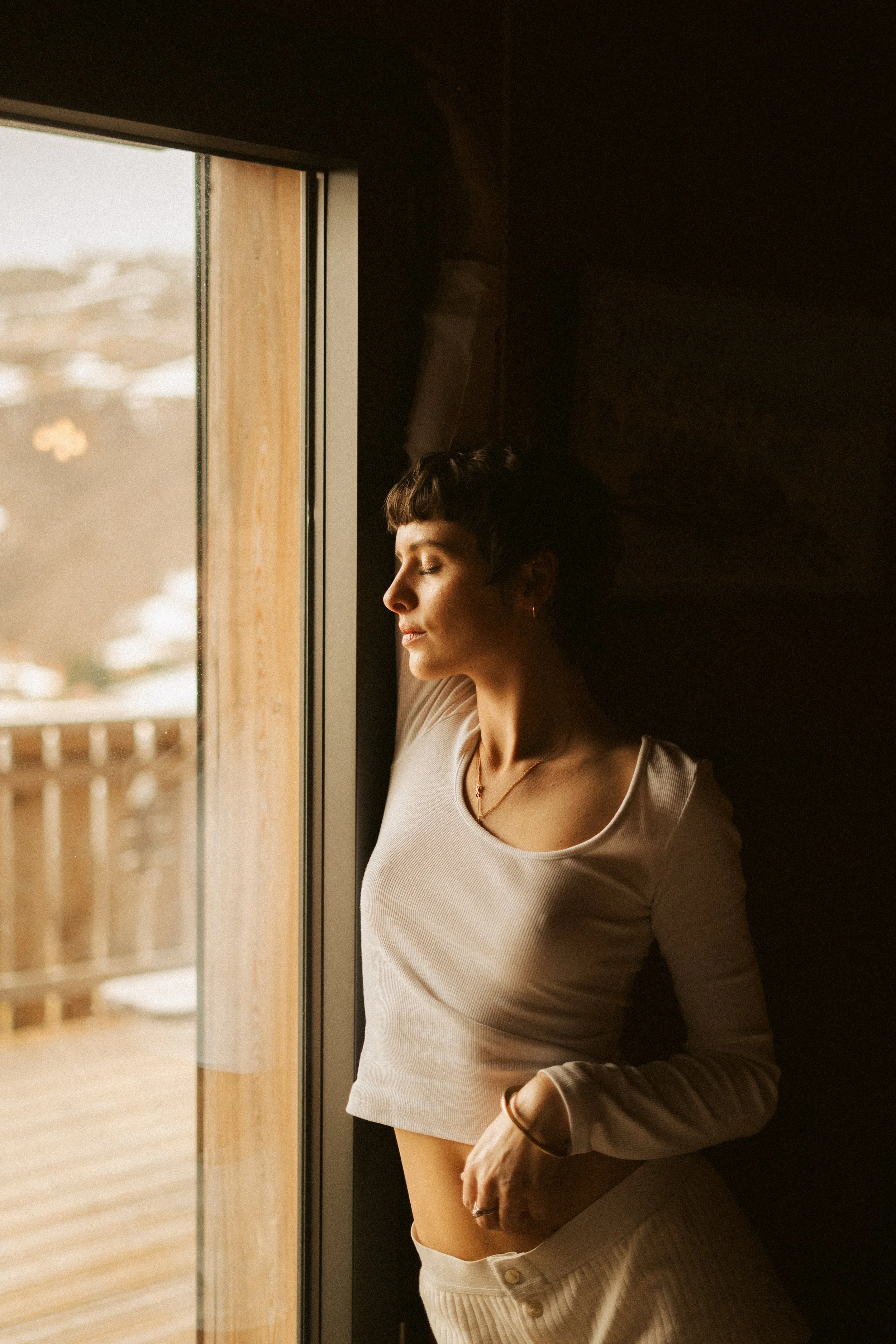 Une femme aux cheveux courts et bouclés, portant un haut moulant blanc, se tient près d'une fenêtre en bois, profitant de la lumière naturelle avec les yeux fermés.
