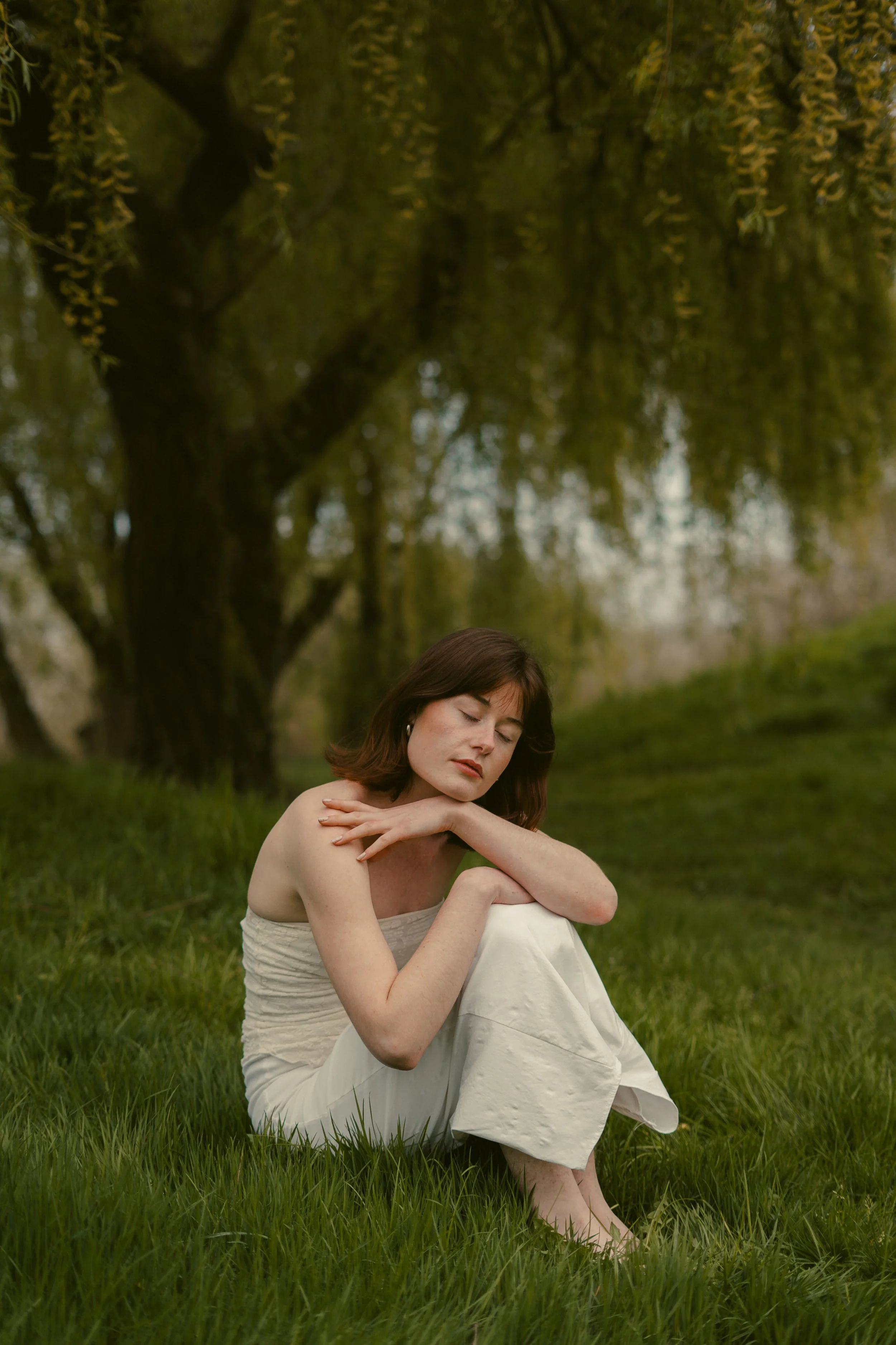 Une femme assise sur l'herbe, les yeux fermés, dans une nature verdoyante, avec un grand arbre en arrière-plan, ambiance calme et rêveuse.