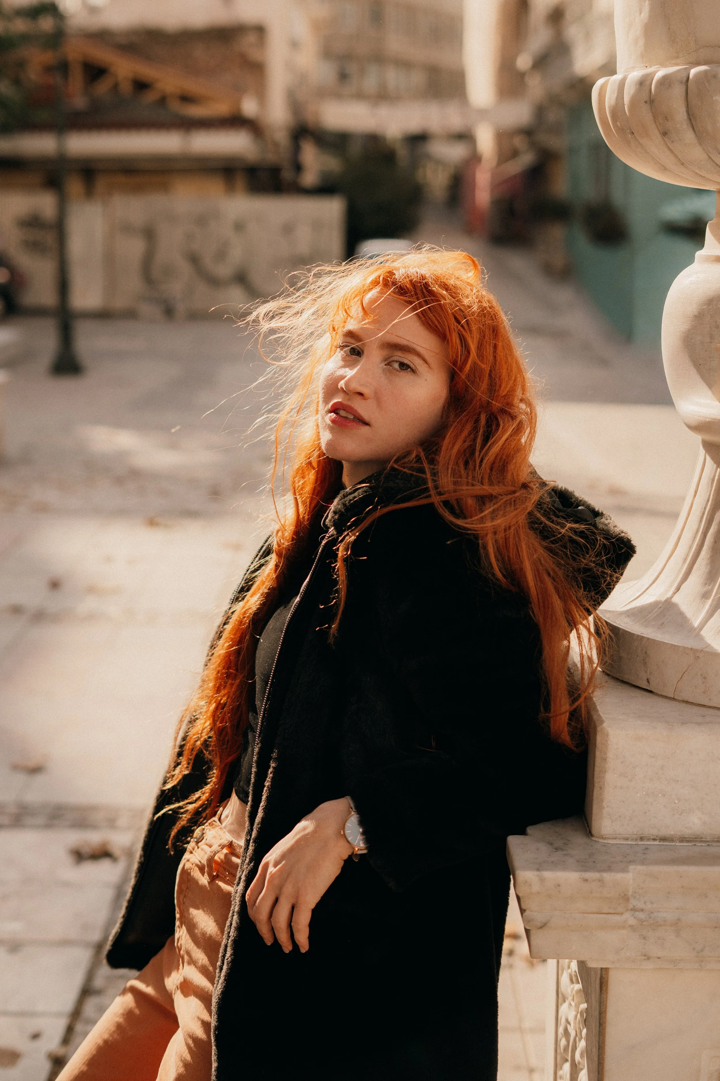 Jeune femme aux cheveux rouges portant un manteau noir, se tenant à côté d'une balustrade en pierre dans un environnement urbain en plein air, éclairé par la lumière naturelle