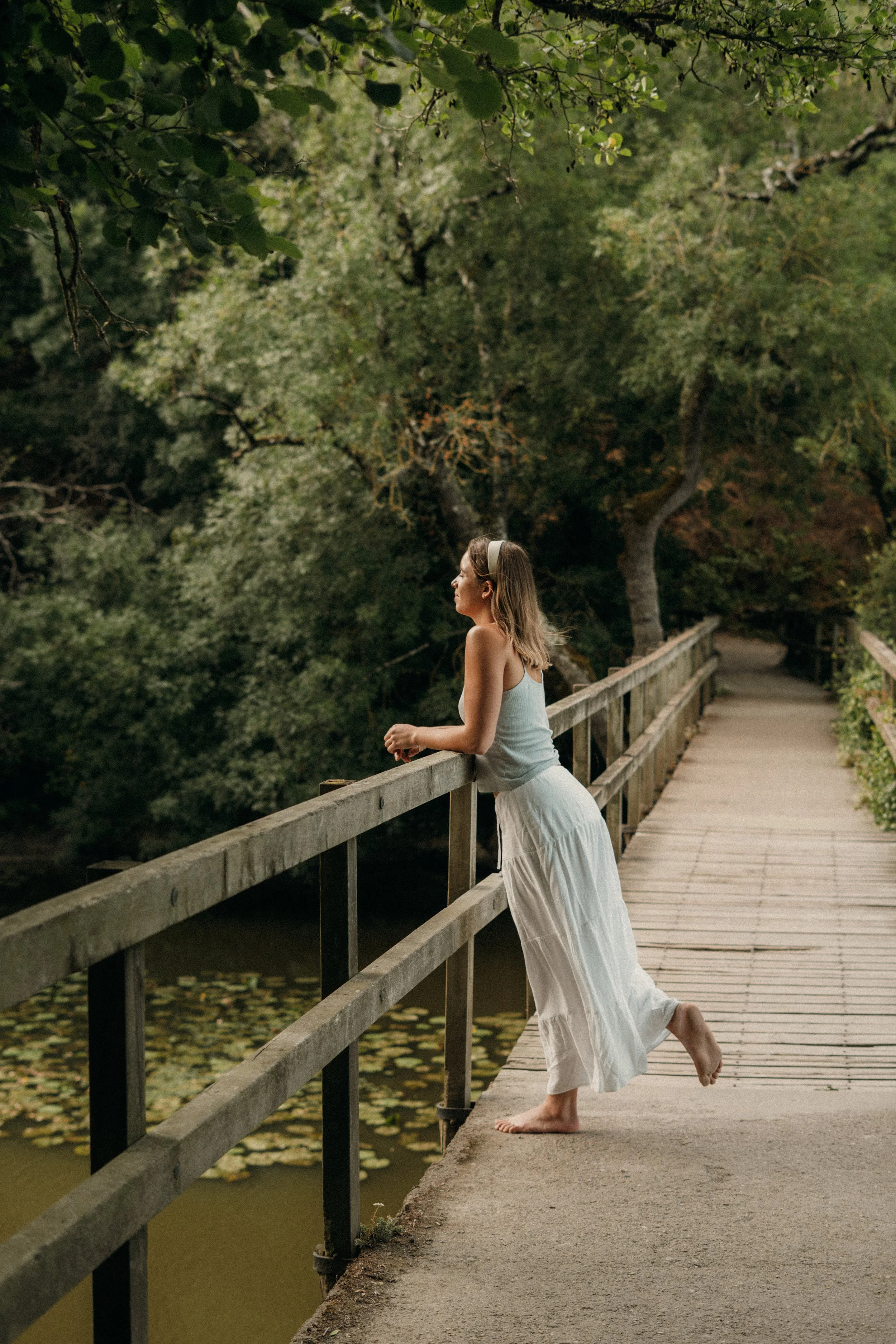 Une femme en robe blanche et bandeau regarde pensivement au-delà de la rambarde sur un pont en bois, entourée d'une forêt verdoyante.