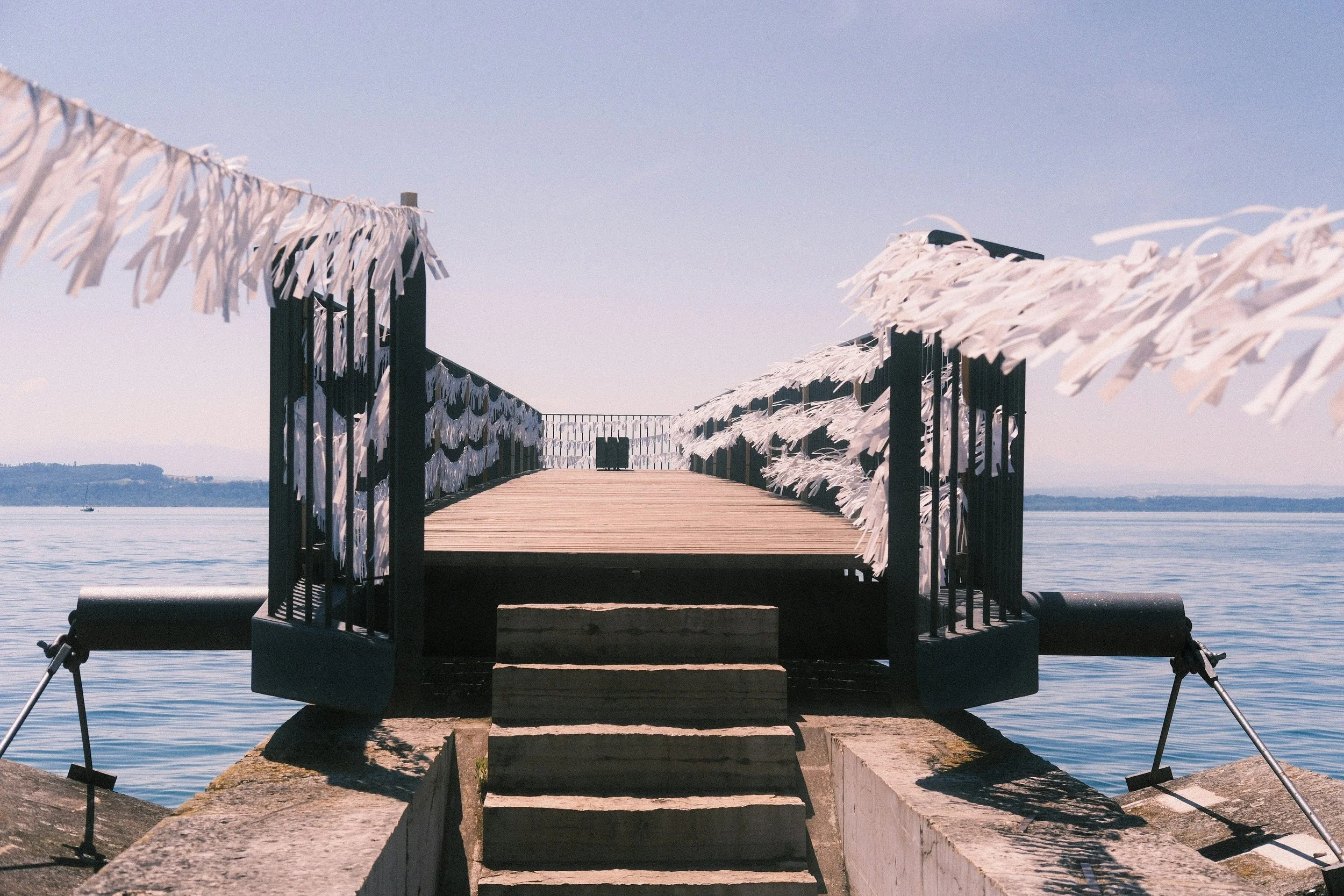 Ein Steg am Wasser mit weißen Stoffbändern an den Geländern, ein paar Steinstufen führen zum Steg, im Hintergrund Wasser und ein Himmel mit wenigen Wolken.