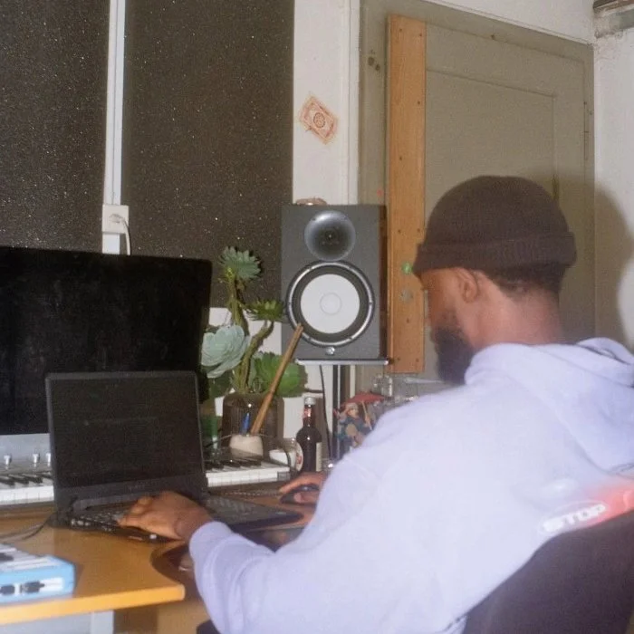 Ein Mann mit dunklem Bart und einer schwarzen Beanie, der an einem Laptop arbeitet, in einem Raum mit einem Computerlautsprecher, Pflanzen, Wasserflaschen und einem schwarzen Monitor.