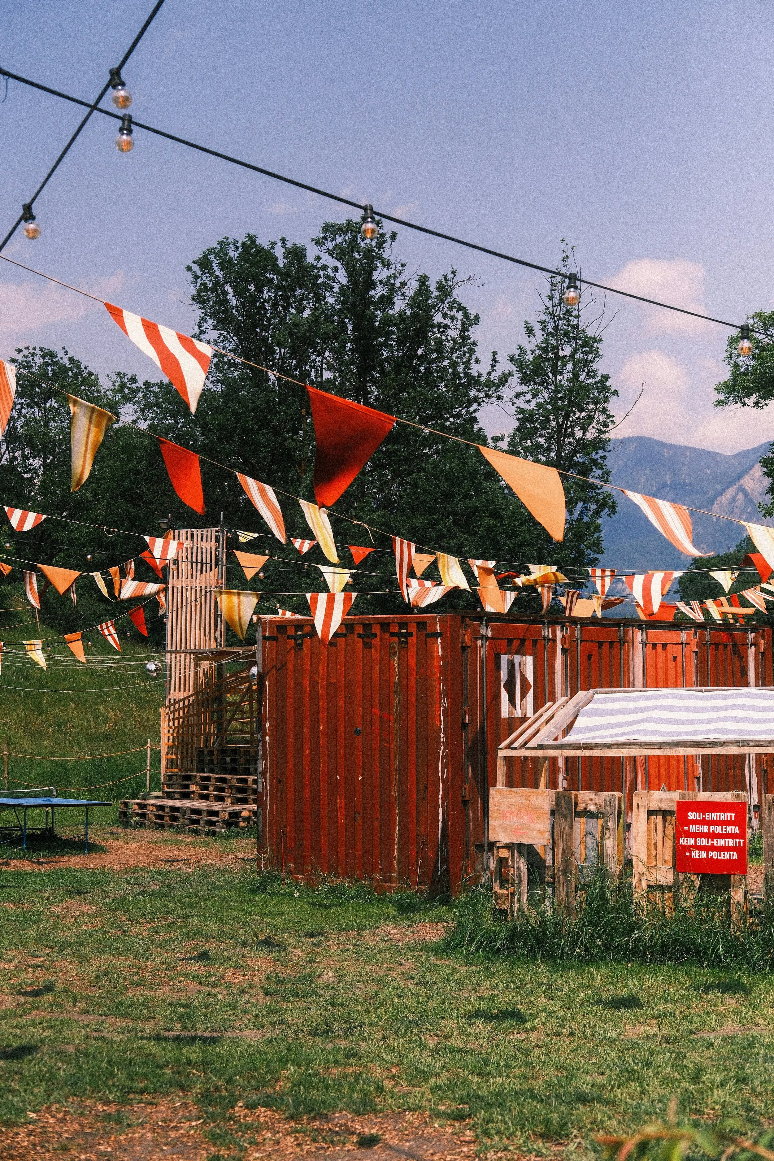 Festlich geschmückter Außenbereich mit bunten Wimpeln und Lichterketten, Holzbarrieren und einem Zelt, mit Bergen im Hintergrund.