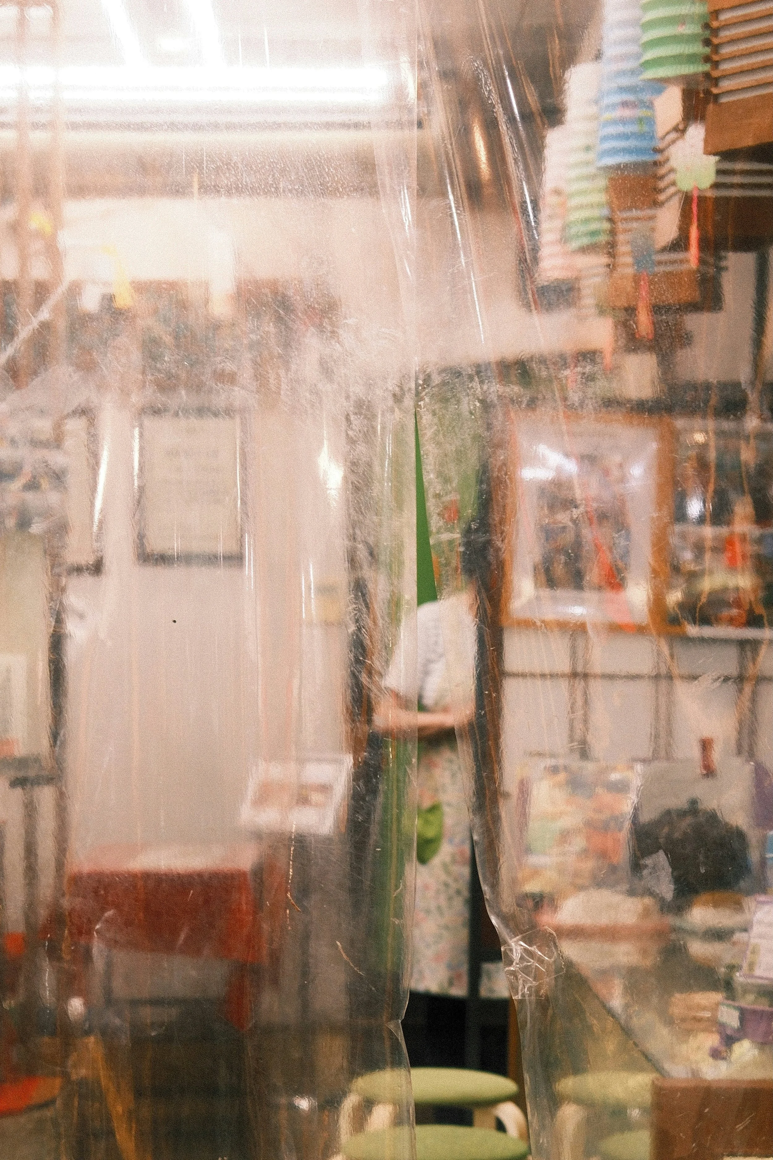 Ein Foto, das durch eine sichtbare, zerknitterte Plastikfolie aufgenommen wurde, mit Blick auf einen Laden im Hintergrund, der verschiedene Produkte und Menschen zeigt.