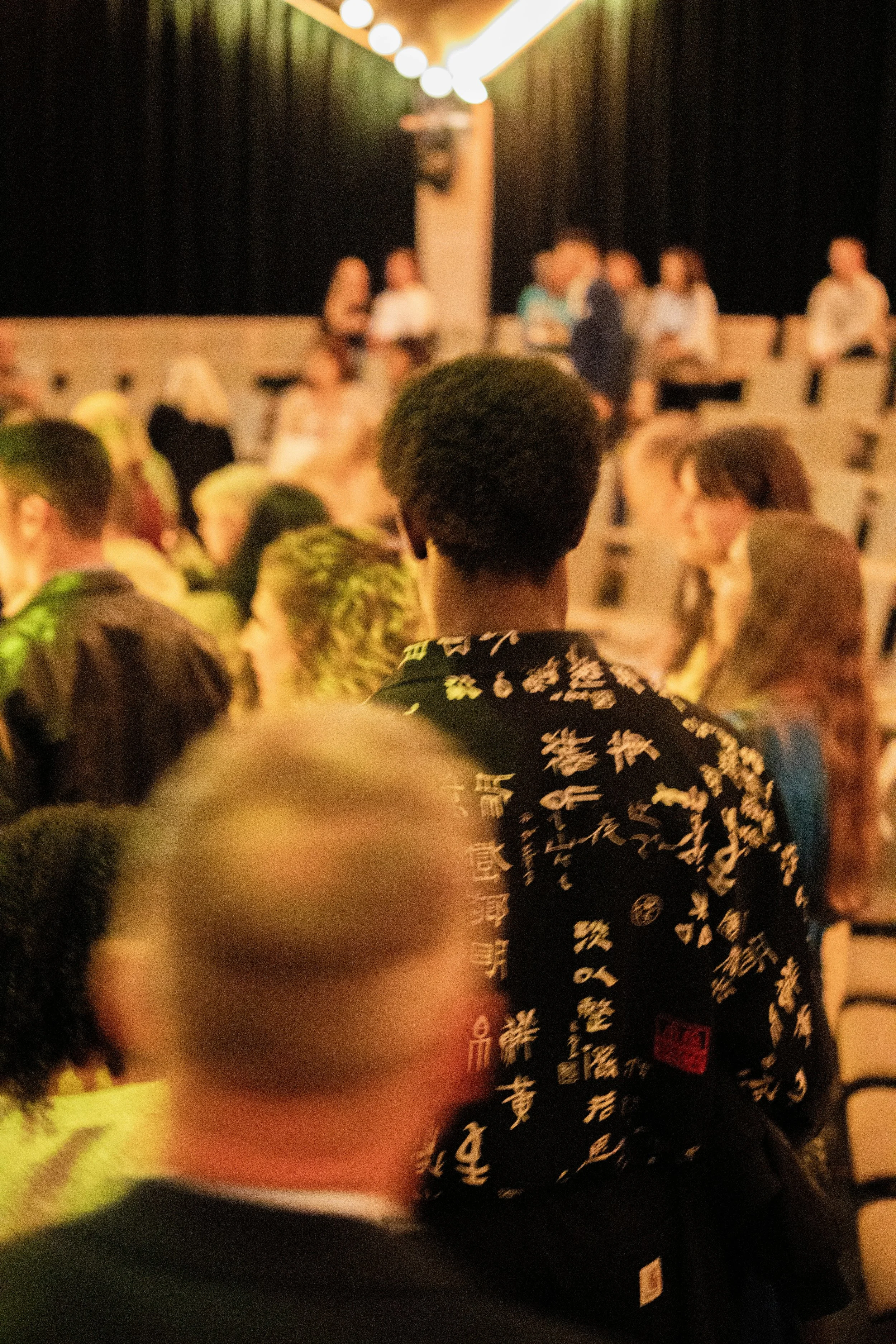 Menschen in einem dunklen Saal, am meisten sichtbar ein junger Mann mit Afro-Haar, der ein kimonoähnliches Kleid mit weißen japanischen Schriftzeichen trägt, im Hintergrund sitzen oder stehen zusätzliche Personen.
