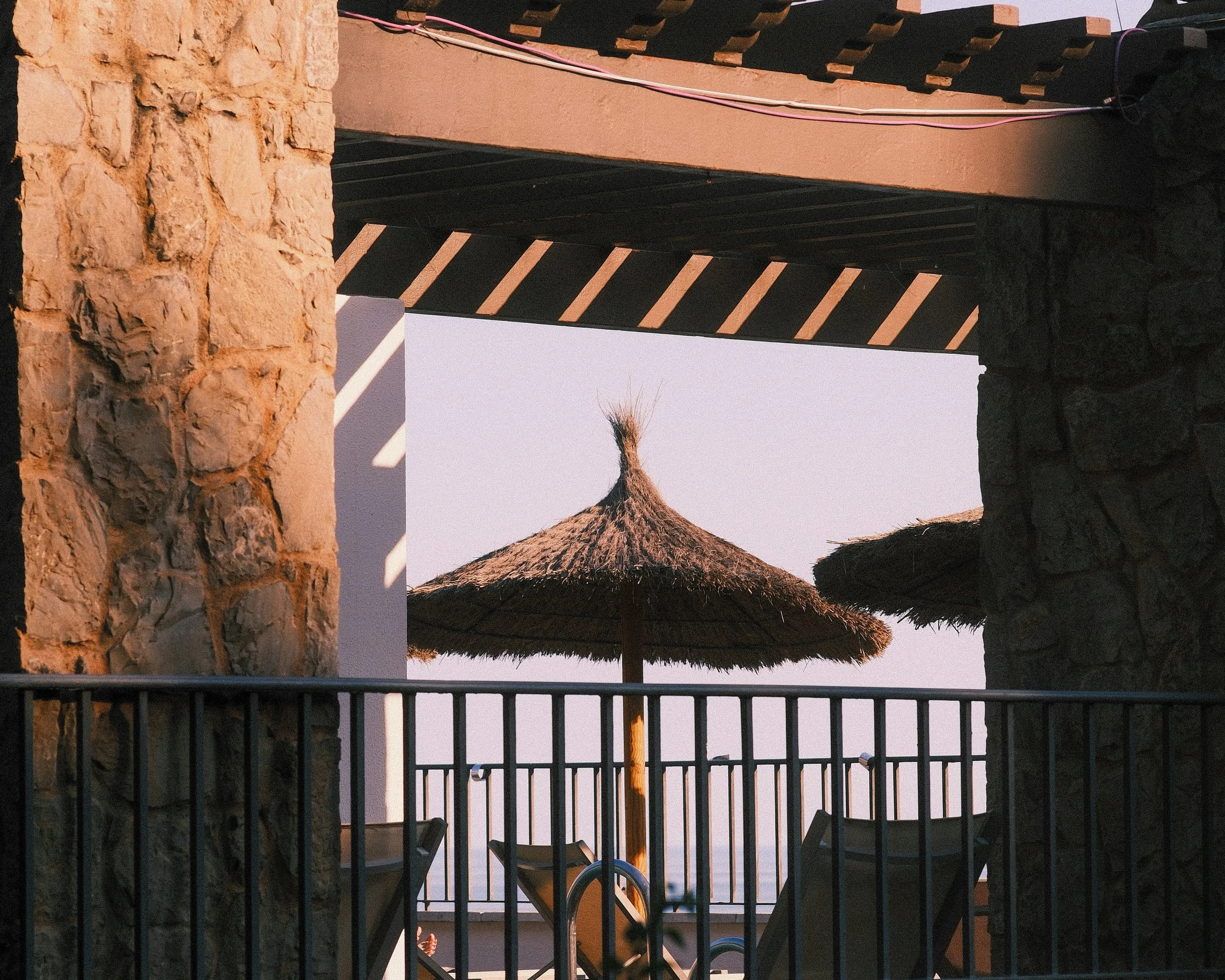 Blick auf einen Strand mit Sonnenschirm und Liegestühlen, gesehen durch eine grillartige Barriere zwischen zwei Steinmauern