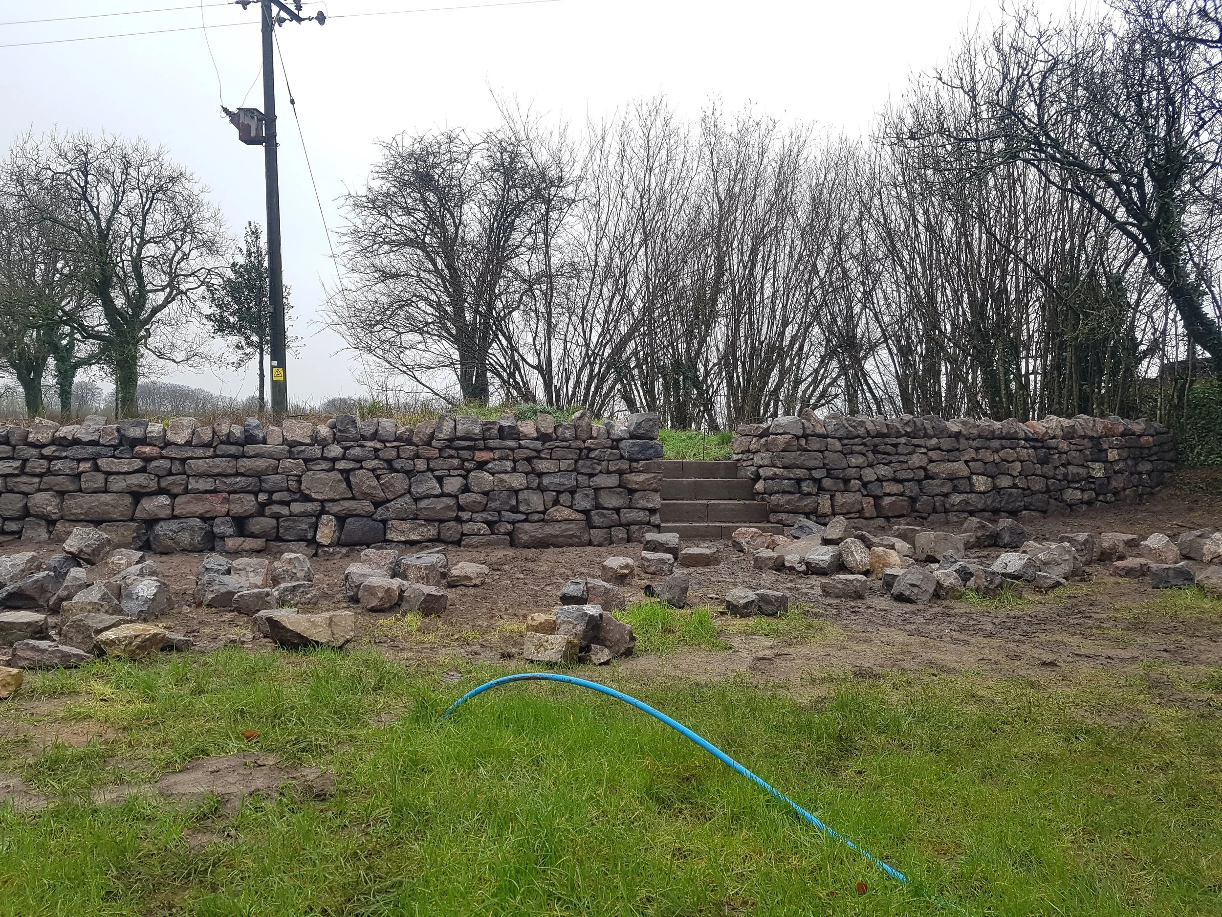 A dry Stone retaining wall with granite steps in Priddy, Somerset. with trees and a utility pole in the background on a cloudy day 